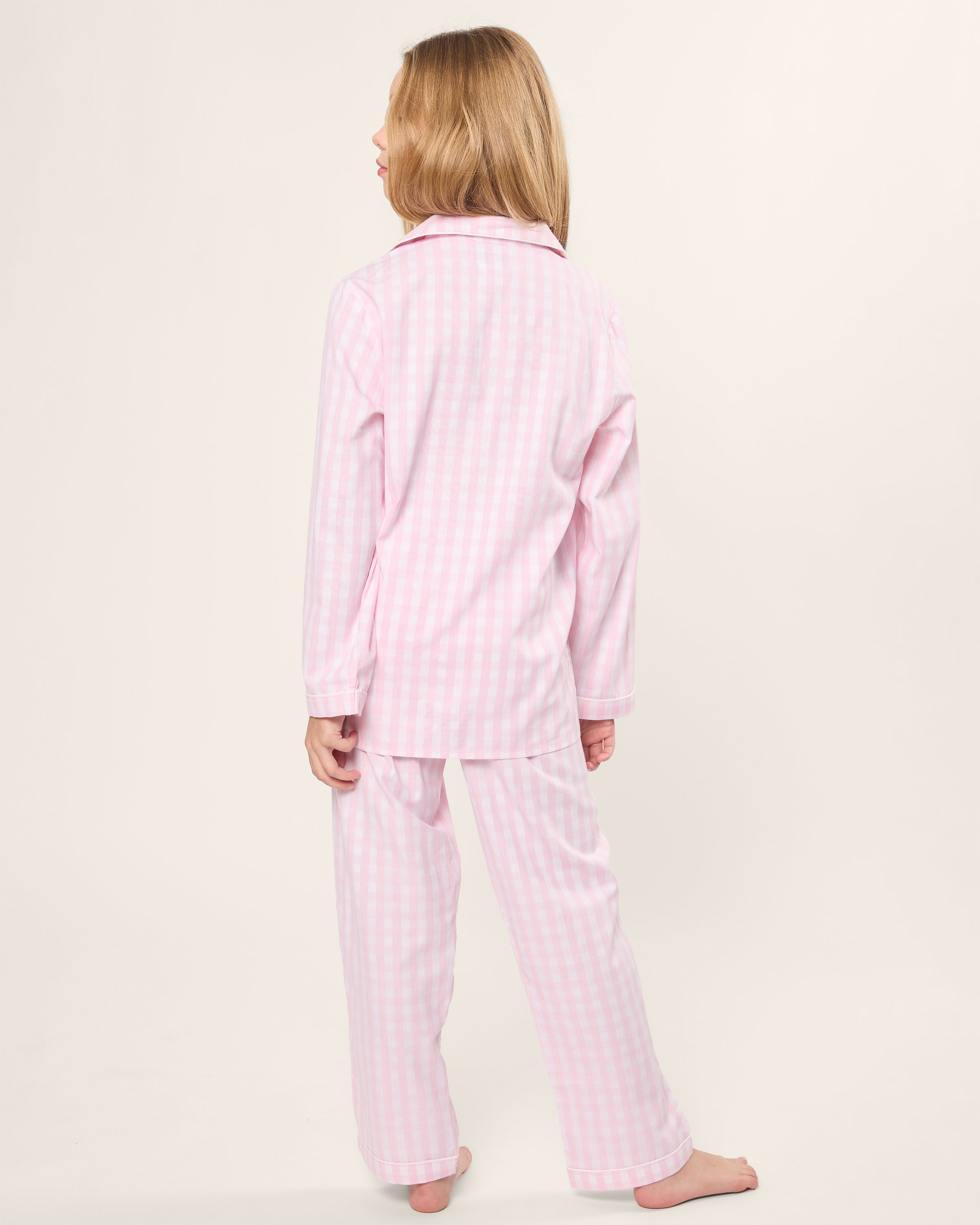 A young girl with shoulder-length light brown hair faces away, barefoot on a plain background, wearing the Petite Plume Kids Twill Pajama Set in Pink Gingham.