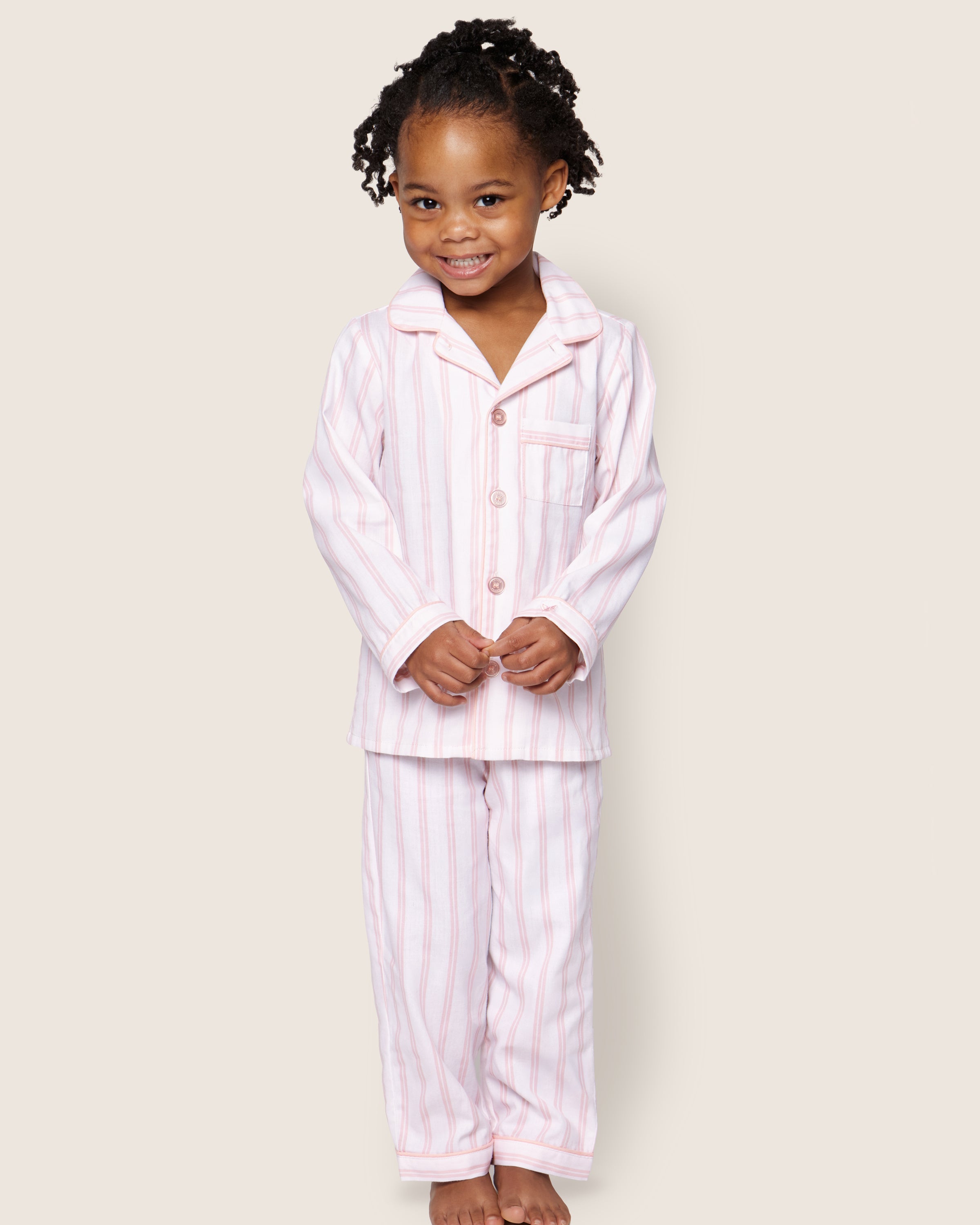 A young child with curly hair smiles while standing barefoot against a plain backdrop, wearing Petite Plumes Kids Twill Pajama Set in Pink and White Stripe, made from luxurious cotton sleepwear with buttons and a pocket.