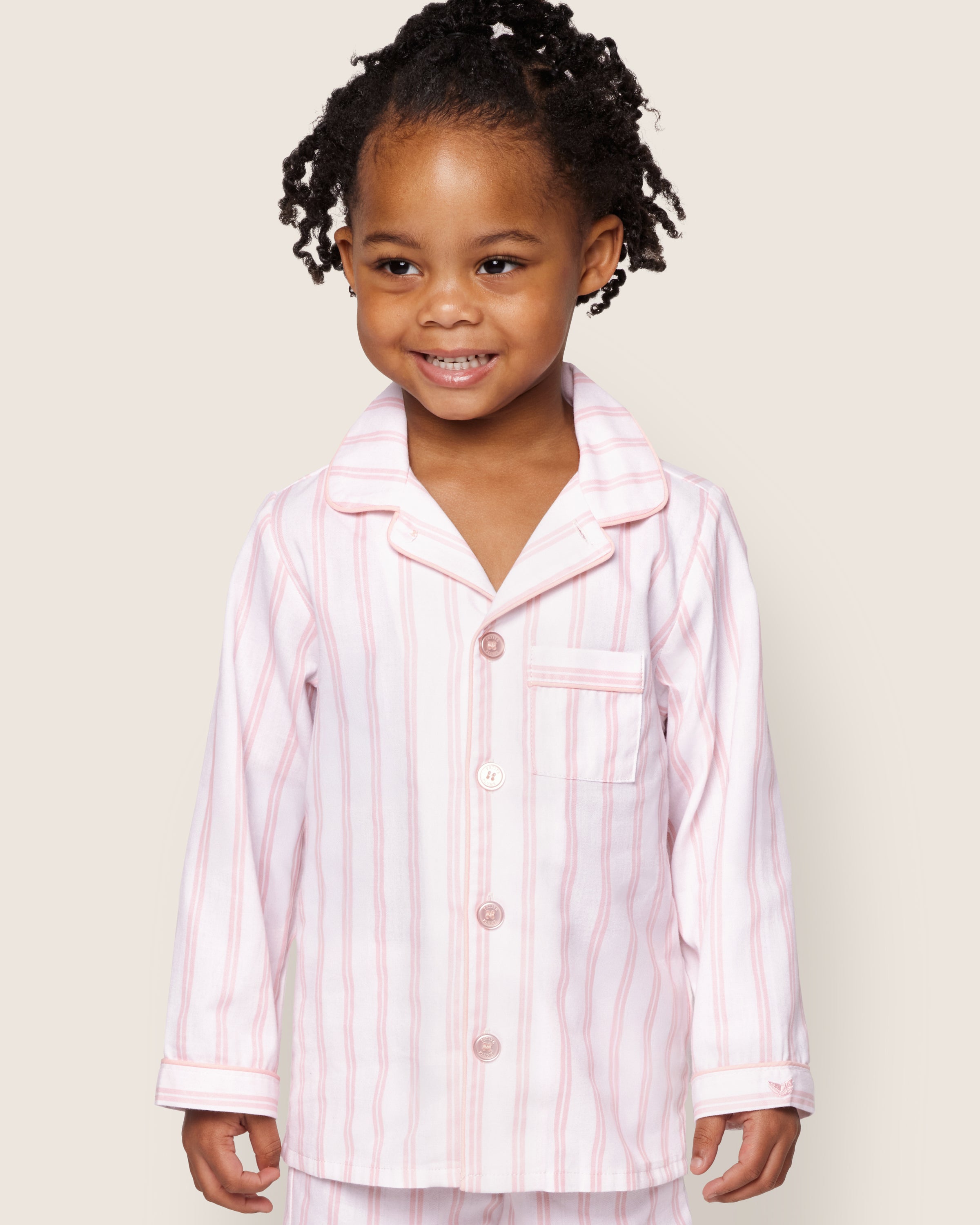 A young child with curly hair smiles while wearing Petite Plumes Kids Twill Pajama Set in Pink and White Stripe, featuring a buttoned top with a pocket on a light cream background.