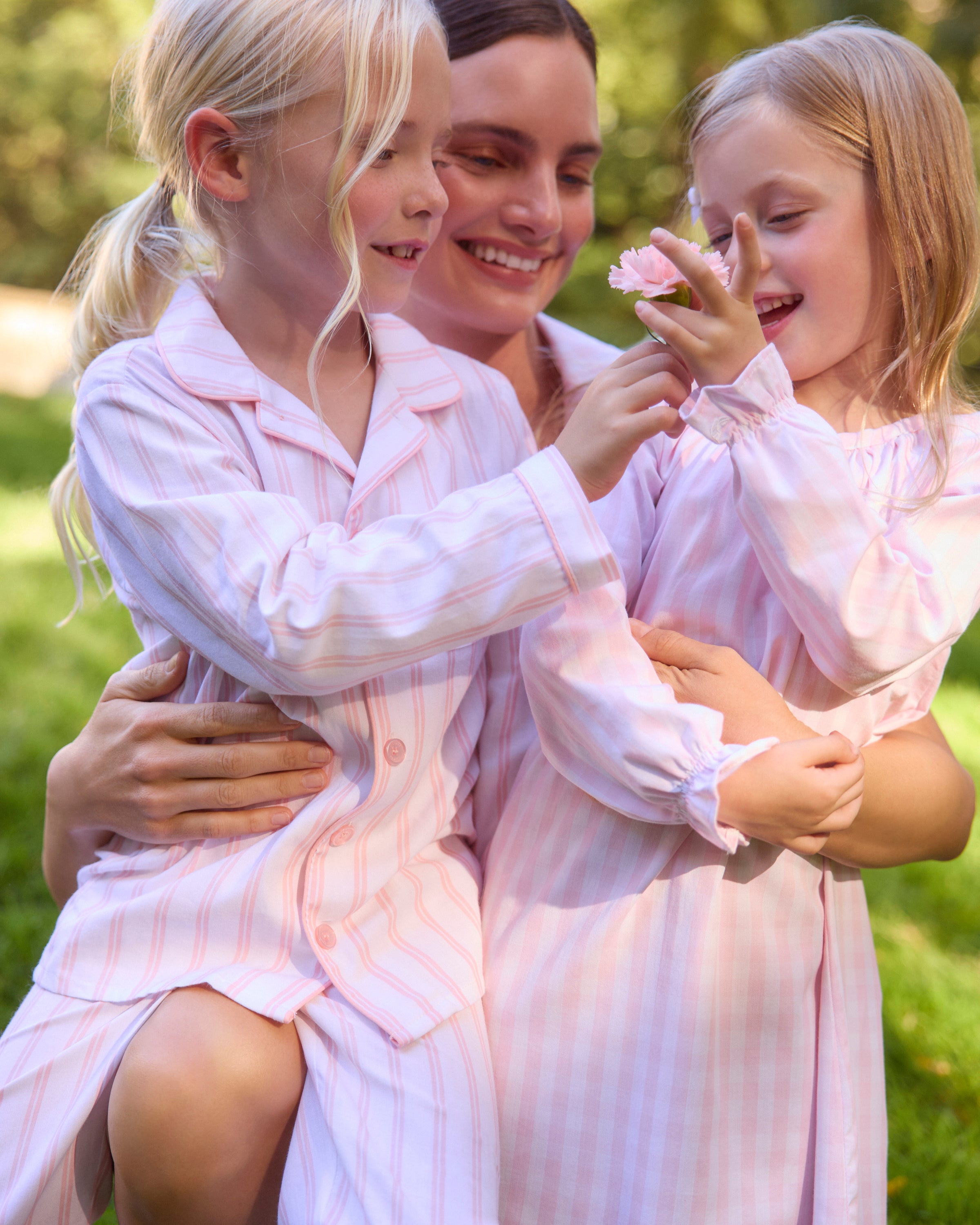 A young child with curly hair smiles while standing barefoot against a plain backdrop, wearing Petite Plumes Kids Twill Pajama Set in Pink and White Stripe, made from luxurious cotton sleepwear with buttons and a pocket.