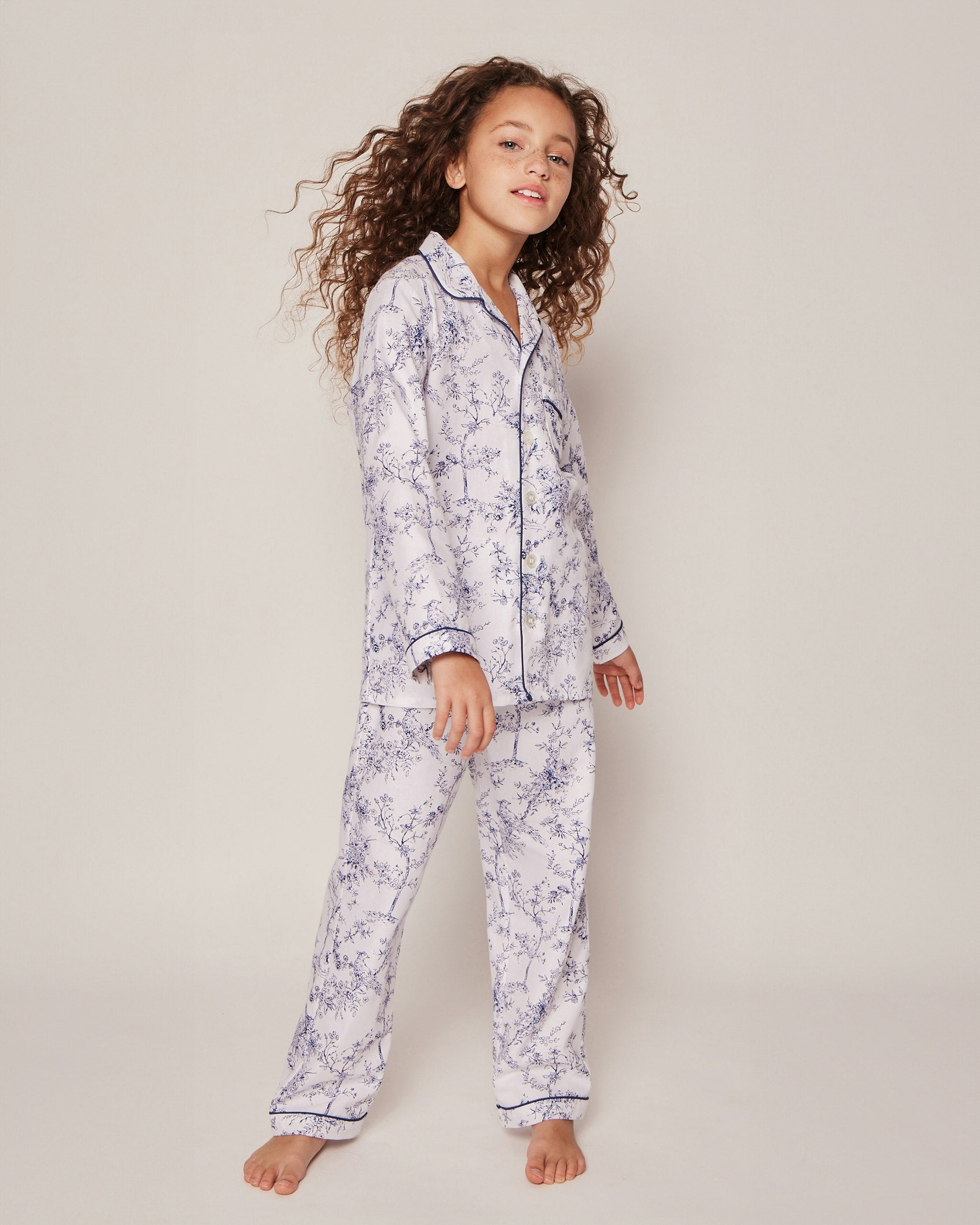 The Kids Twill Pajama Set in Timeless Toile by Petite Plume features a long-sleeve, luxury sleepwear style with a classic collar and matching pants. Its white background showcases an intricate blue floral and bird pattern for a vintage, sophisticated charm.