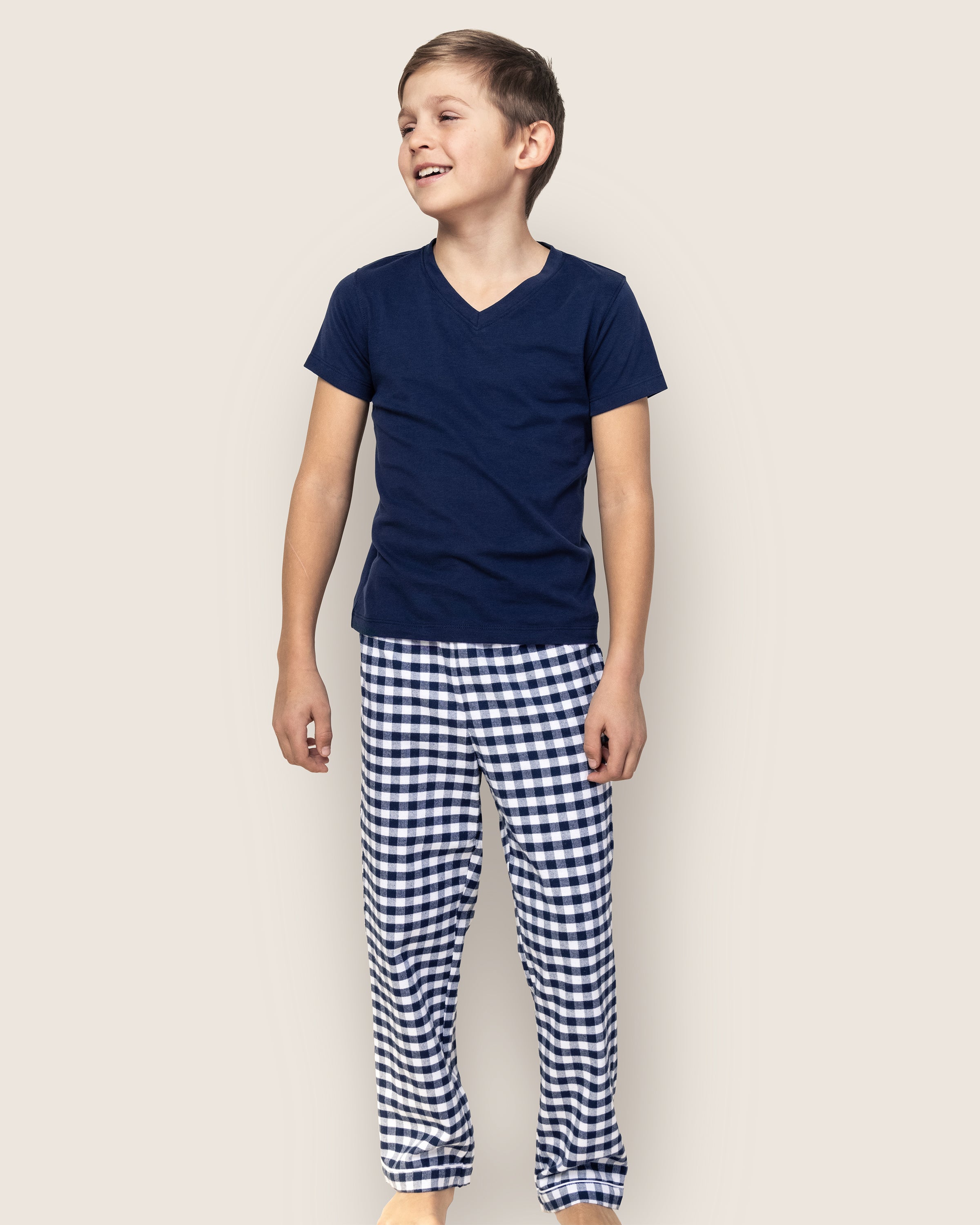 A smiling child wearing a navy blue t-shirt and Petite Plumes Kids Twill Pajama Pants in Navy Gingham stands against a plain background.