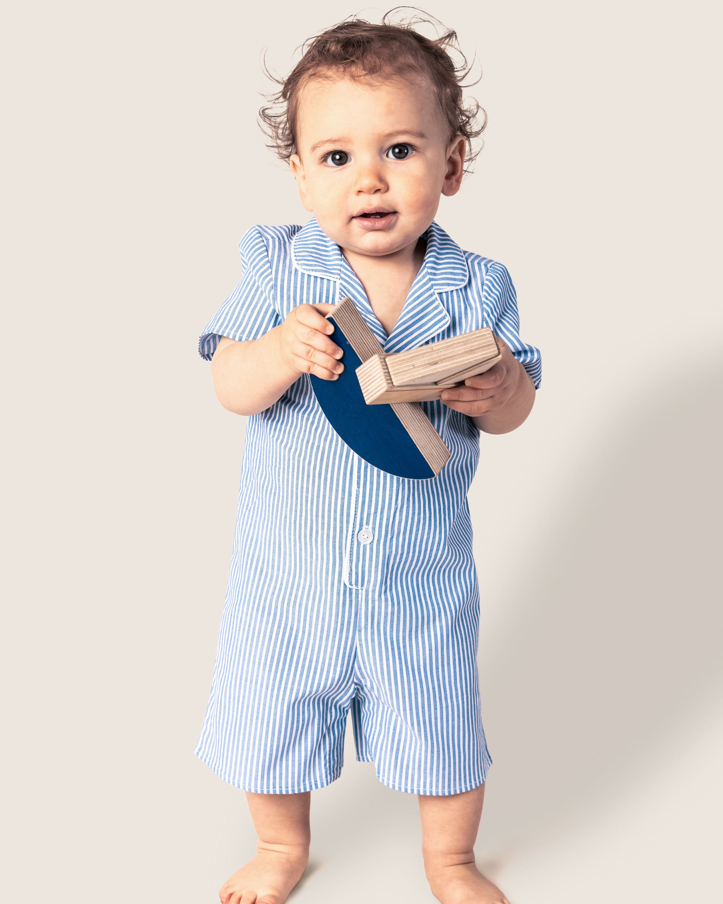 A toddler wearing Petite Plumes Babys Twill Summer Romper in French Blue Seersucker stands barefoot, holding blue and plain wooden blocks. Against a light beige backdrop, the child’s curly hair and curious expression are highlighted.