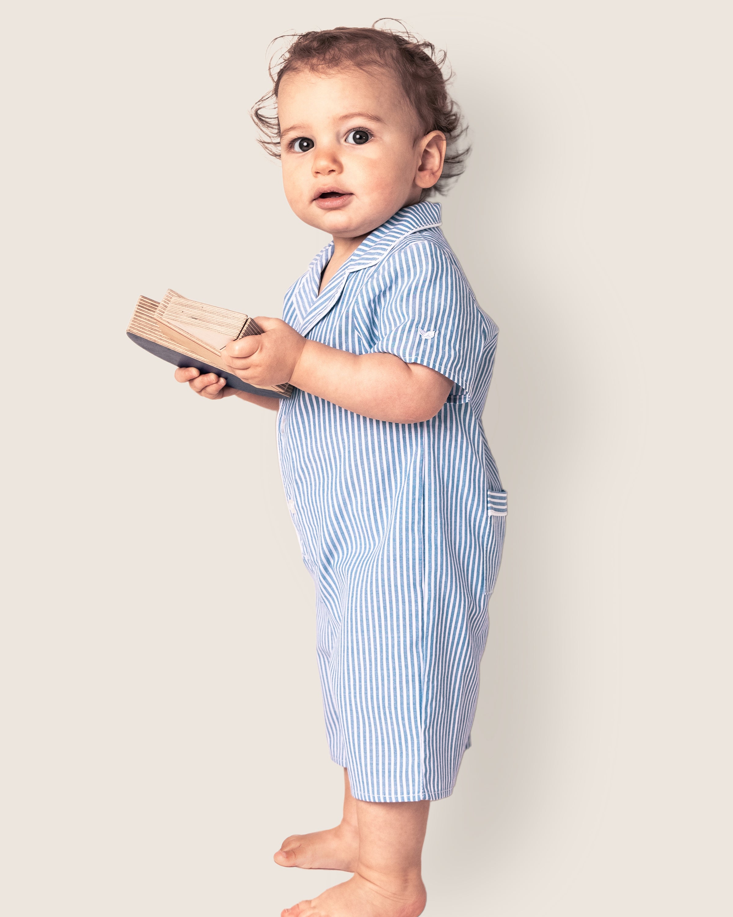 A curly-haired toddler stands barefoot with a small book, dressed in Petite Plumes Babys Twill Summer Romper in French Blue Seersucker, gazing upwards against an off-white background.