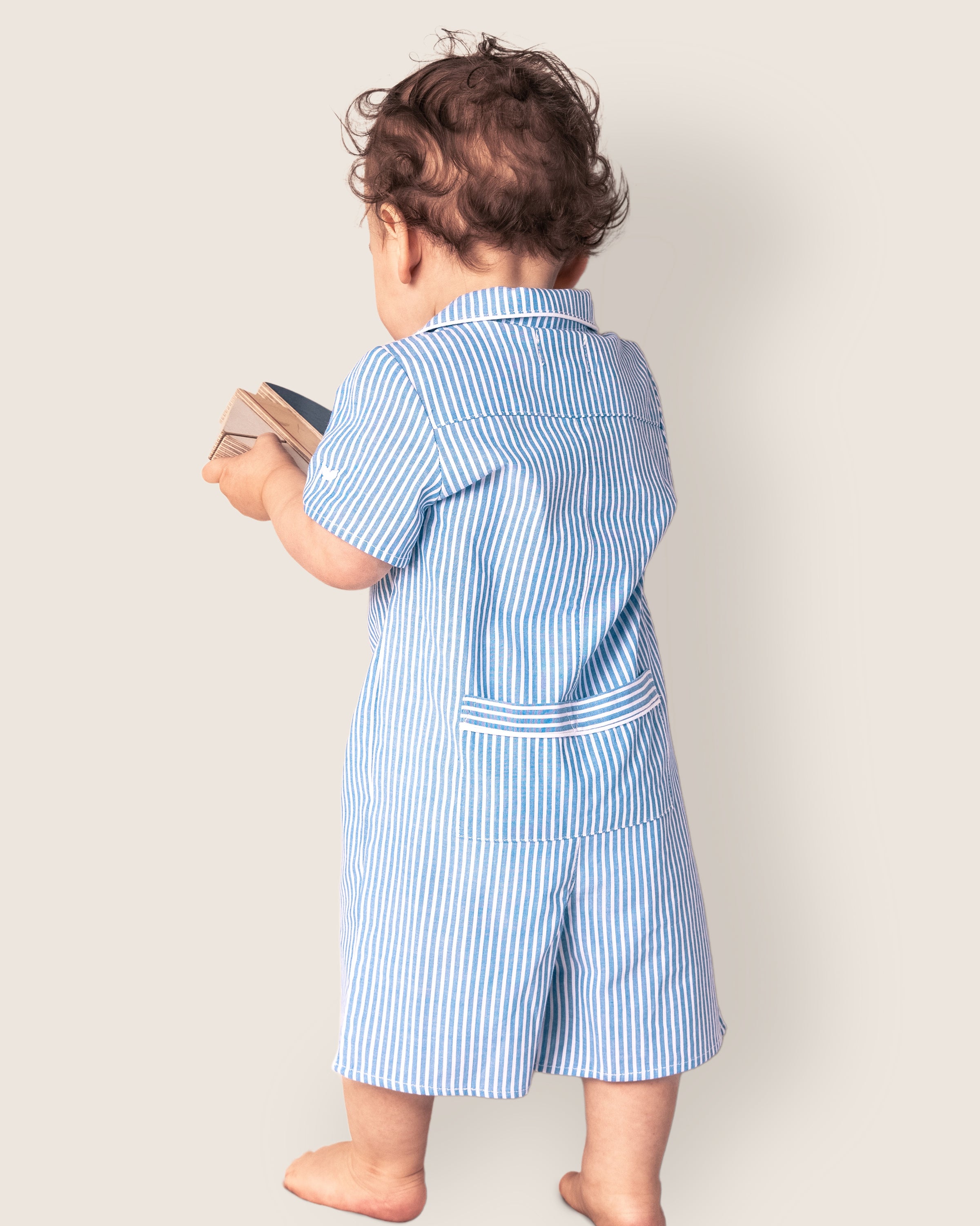 A toddler with curly hair stands barefoot, facing away in a Petite Plume Babys Twill Summer Romper in French Blue Seersucker, clutching a book against a light background that accentuates their innocence and charm.