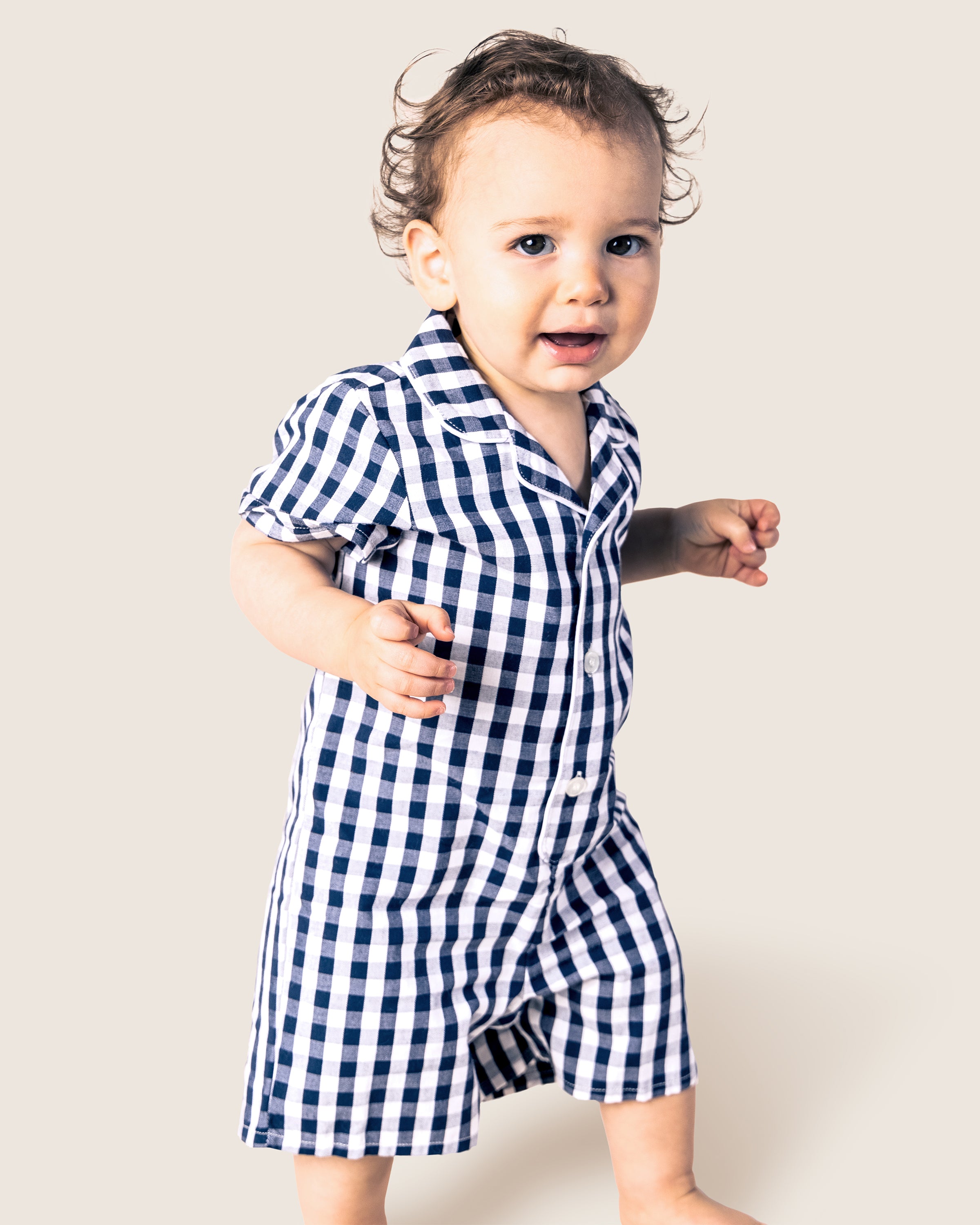 A young child wearing Petite Plumes Babys Twill Summer Romper in Navy Gingham smiles, mid-step, against a light background with curly, tousled hair and one arm bent while the other is slightly extended.