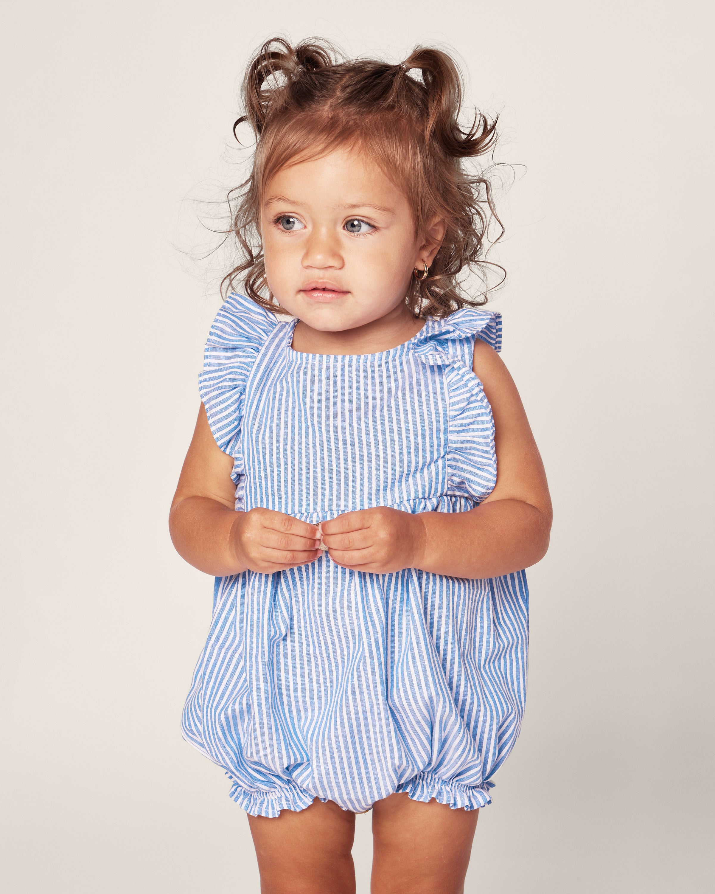 A young child with curly hair wears a Petite Plume Babys Twill Ruffled Romper in French Blue Seersucker with blue and white stripes. She clasps her hands, gazing to the side against a plain light background.