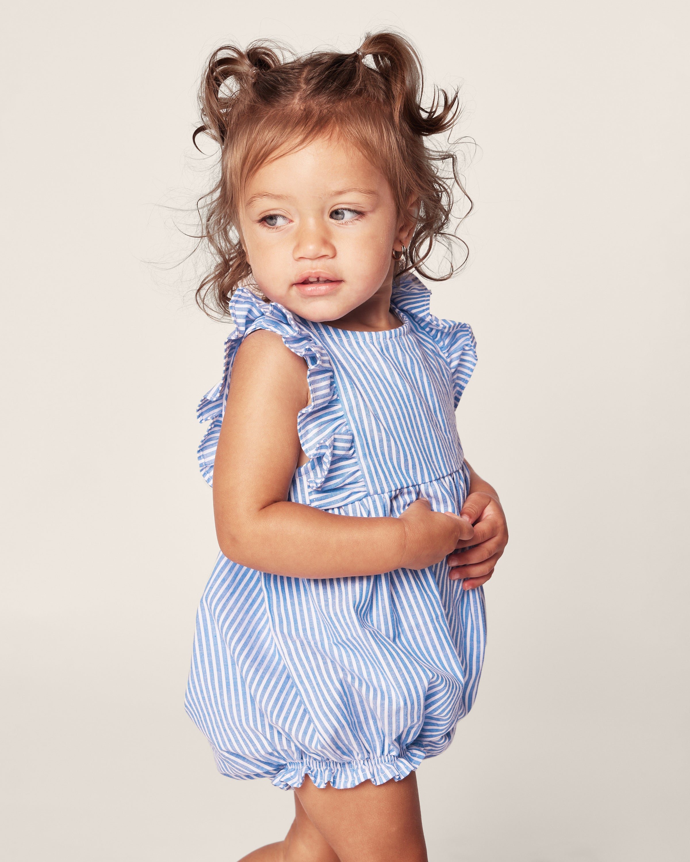 A young girl with brown pigtails wears the Petite Plume Babys Twill Ruffled Romper in French Blue Seersucker, with light blue and white stripes and ruffled shoulders. She gazes to the side, looking relaxed against a plain background.