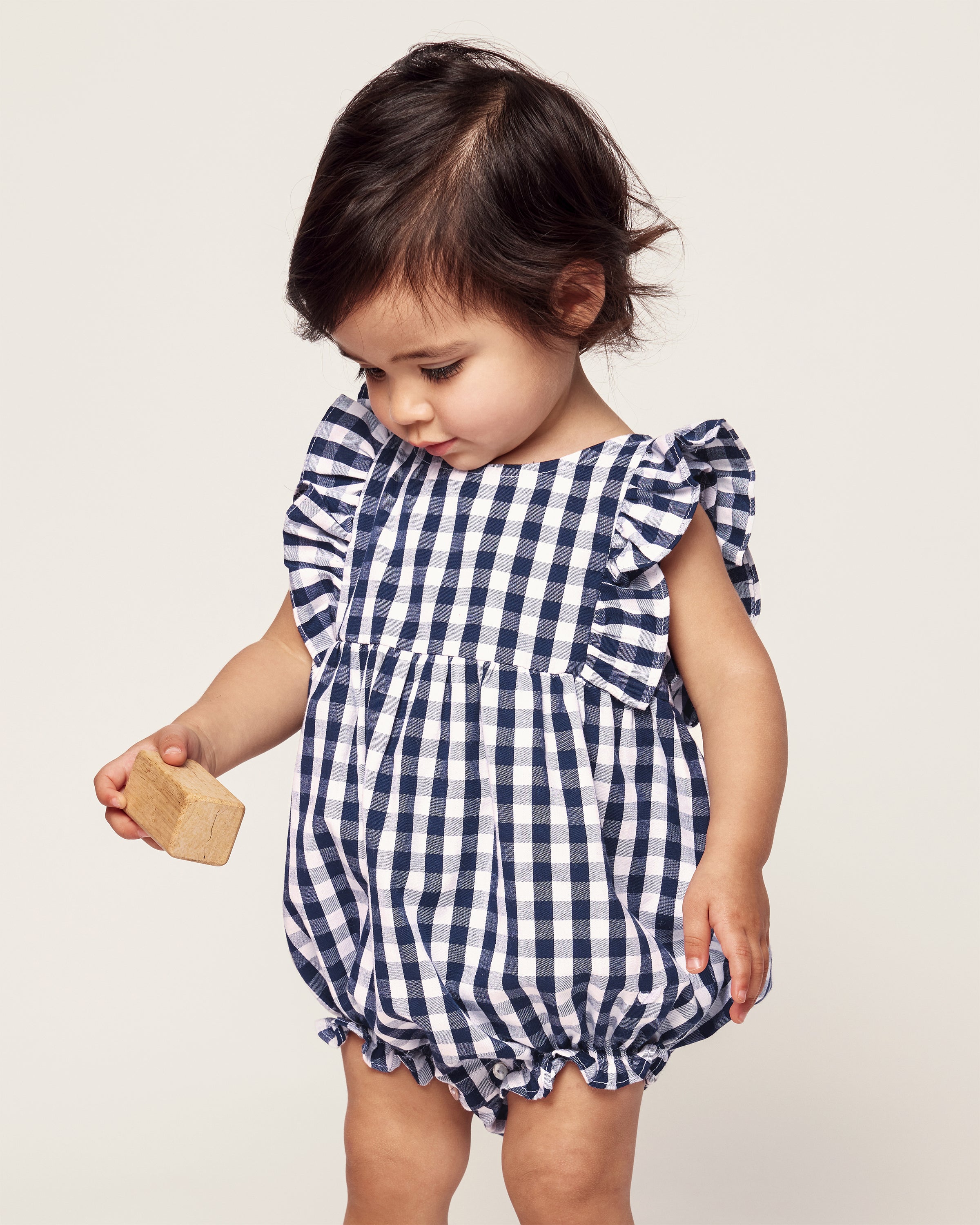 A toddler wearing Petite Plumes Babys Twill Ruffled Romper in Navy Gingham holds a small wooden block. With short dark hair, the child looks downwards against a light, neutral background.