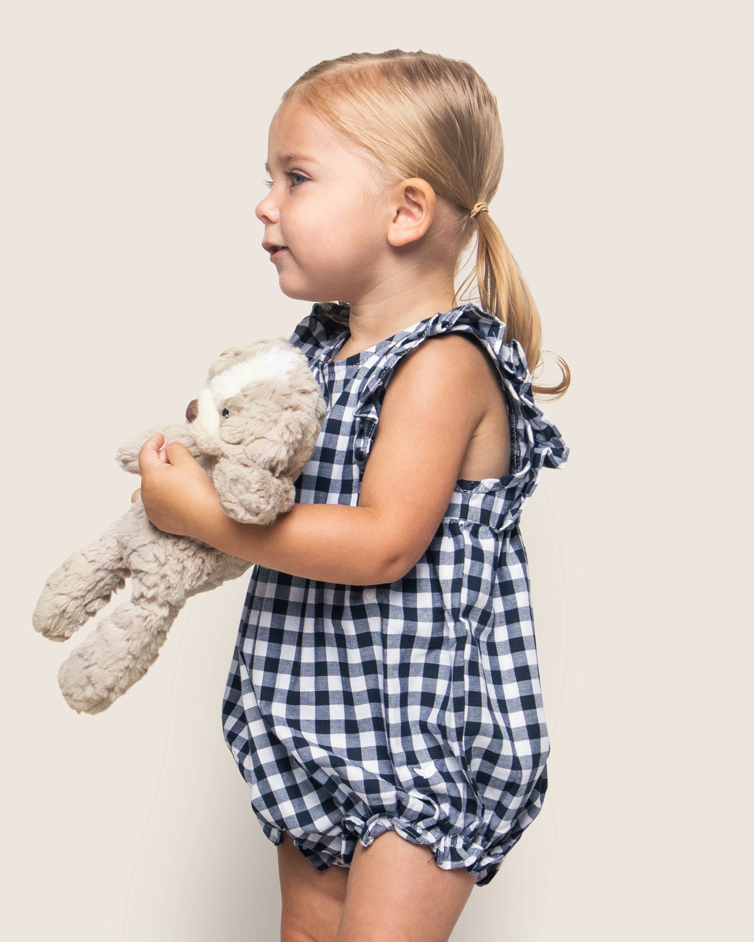 A young child with a blonde ponytail wears Petite Plumes charming Babys Twill Ruffled Romper in Navy Gingham, ideal for sleepwear, while holding a plush toy dog and gazing sideways against a plain light background.