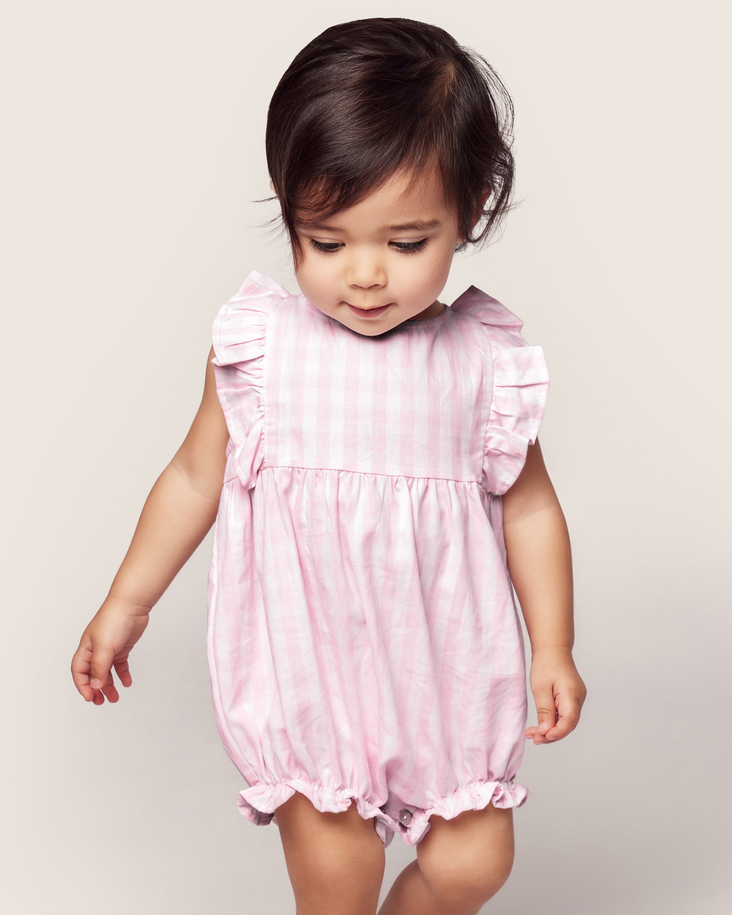 A toddler with dark hair stands against a plain background in Petite Plumes Babys Twill Ruffled Romper in Pink Gingham, featuring ruffled sleeves, as they look down with a gentle expression, enhancing the rompers charming design.