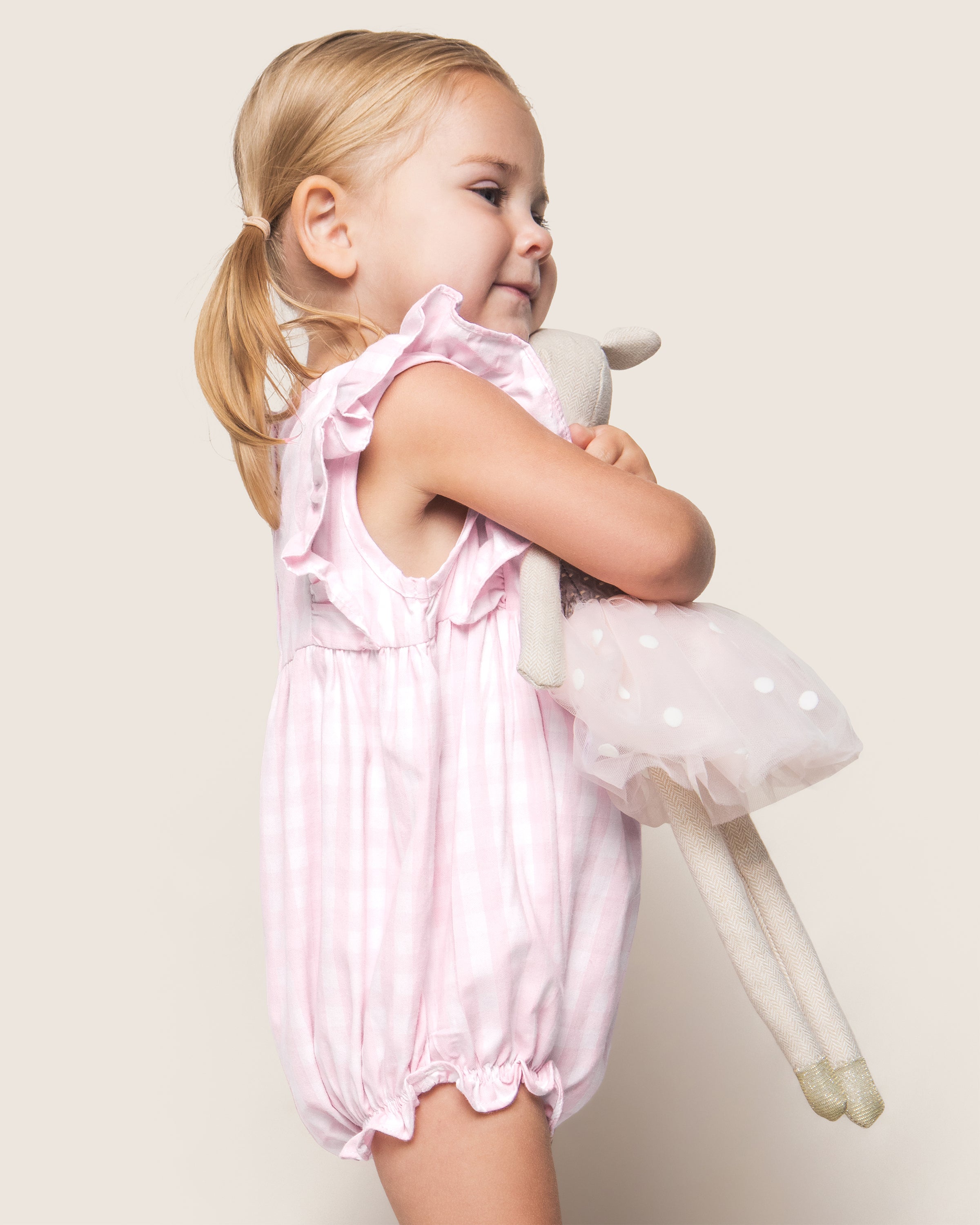 A young child with blonde pigtails beams while wearing Petite Plumes Baby’s Twill Ruffled Romper in Pink Gingham. Her romper, made of flame-retardant fabric, features charming ruffled sleeves as she hugs a stuffed animal in a polka dot dress against a plain, light background.