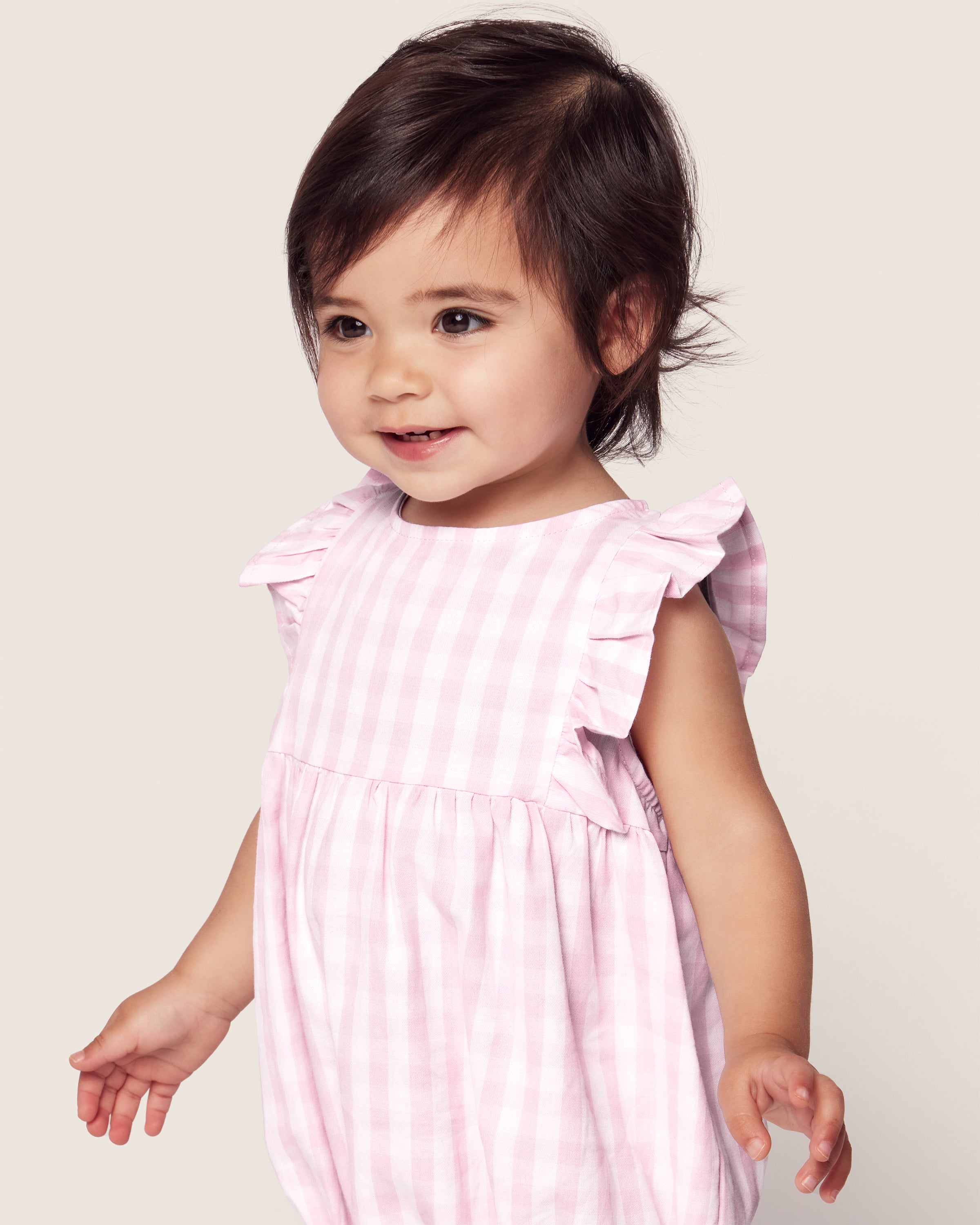 A toddler wearing Petite Plumes Babys Twill Ruffled Romper in Pink Gingham stands smiling against a plain background, with dark hair styled in a short, neat cut.