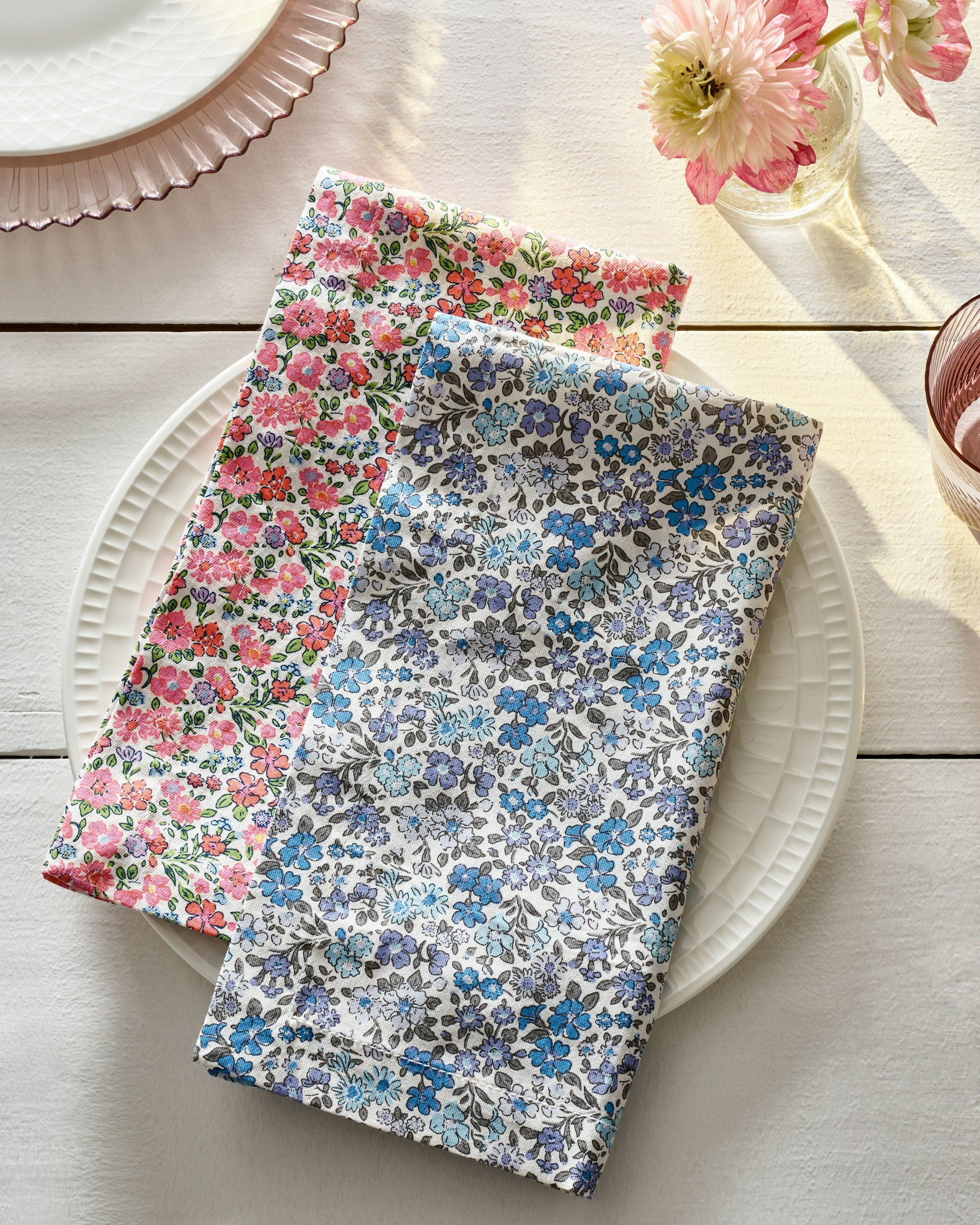 Two Signature Twill Napkins in Fleur DAzur by Petite Plume, adorned with pink and blue floral patterns, rest neatly folded on a white plate. Crafted from 100% cotton, these linens sit on a light wooden table beside a vase of pink flowers and another plates edge as sunlight casts soft shadows.