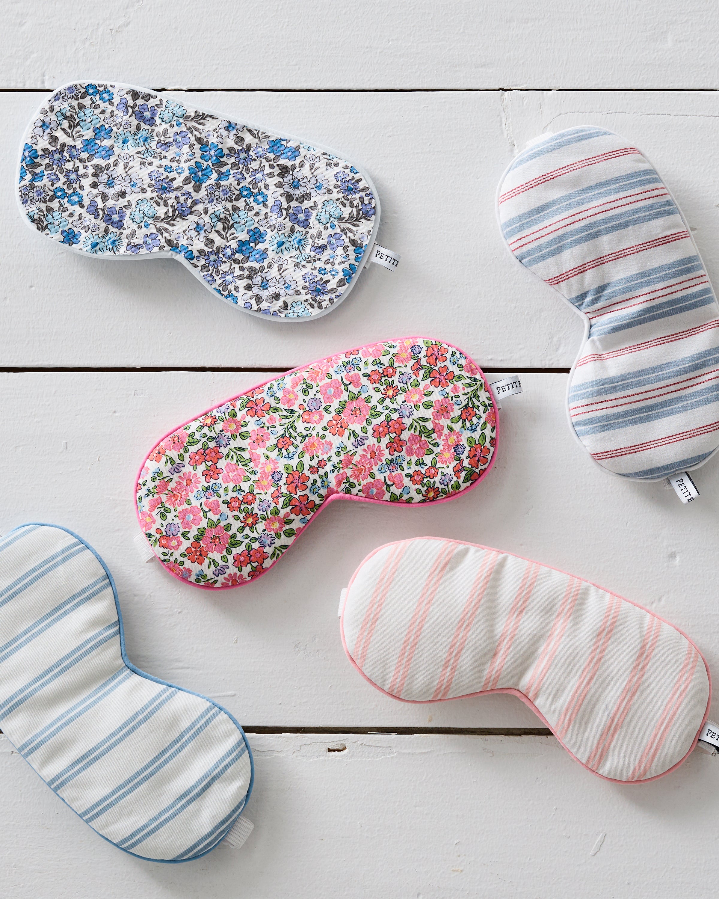 Five luxurious Petite Plume eye masks, elegantly arranged on a white wooden surface, include two in periwinkle stripes and three with floral designs in blue, pink, and red. Perfectly monogrammable for the stylish sleeper.