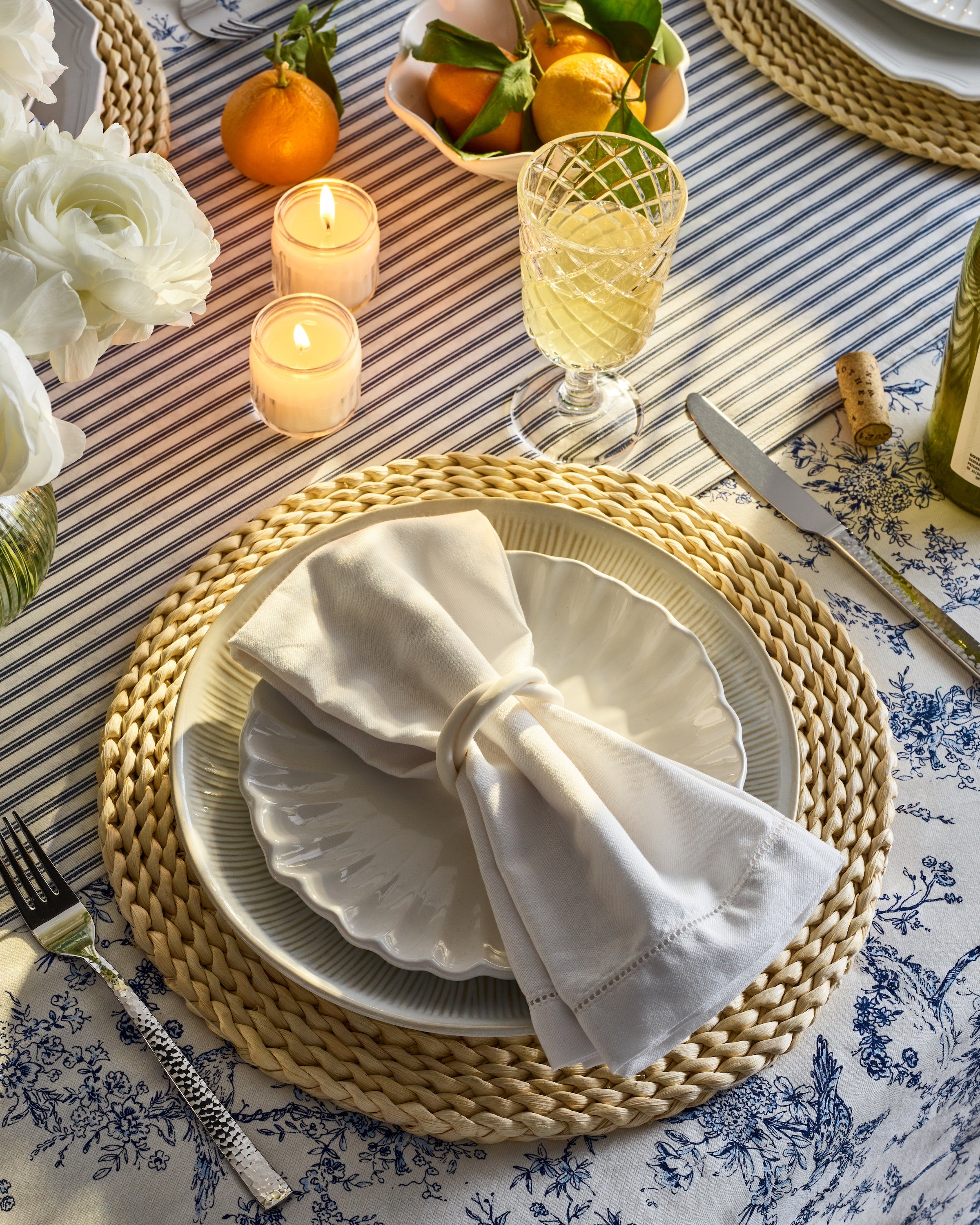 A festive tablescape includes white plates on a woven placemat, topped with Petite Plumes Signature Twill Napkins in White, accompanied by a wine glass and candles. Oranges with leaves rest on linens featuring blue floral patterns, surrounded by delicate white flowers.