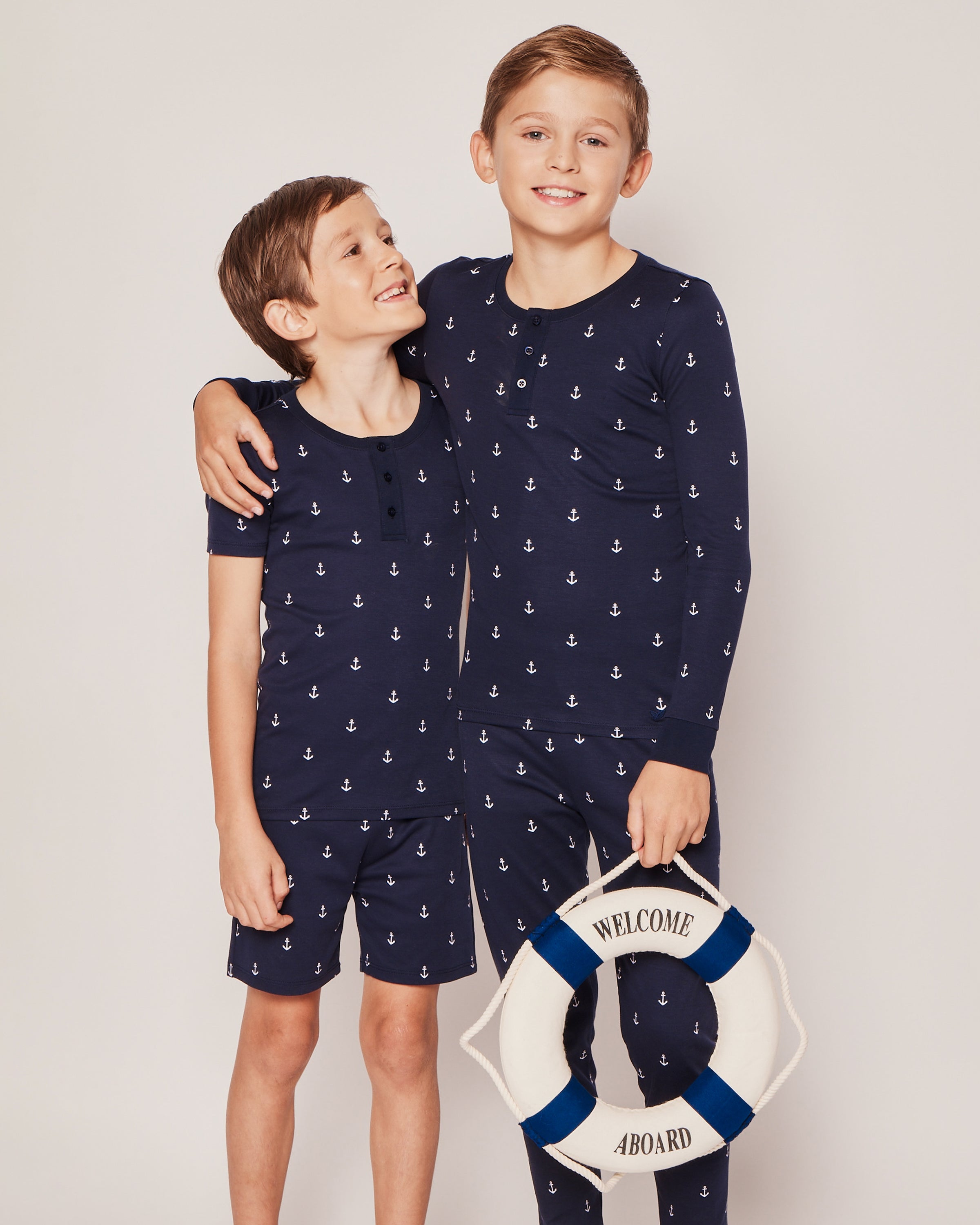 Two boys in matching Petite Plume Kids Pima Snug Fit Pajama Set in Portsmouth Anchors, made from soft Pima Cotton. The older boy holds a life preserver with Welcome Aboard on it. Theyre smiling, and the younger one admires the older in their cozy sleepwear.