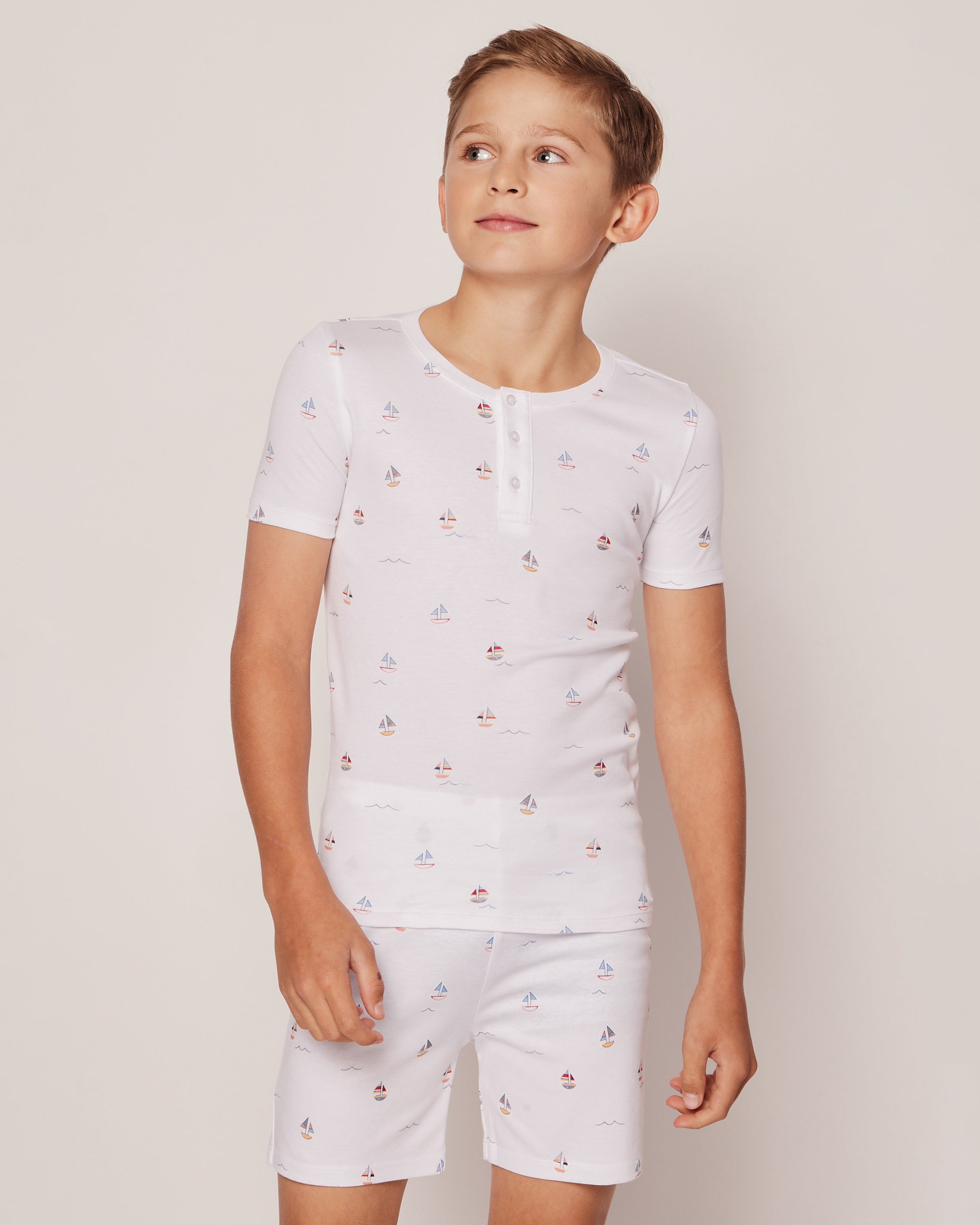A boy wears the Kids Pima Snug Fit Pajama Short Set in Bateau by Petite Plume, featuring colorful prints, as he stands against a plain background, gazing to the side.