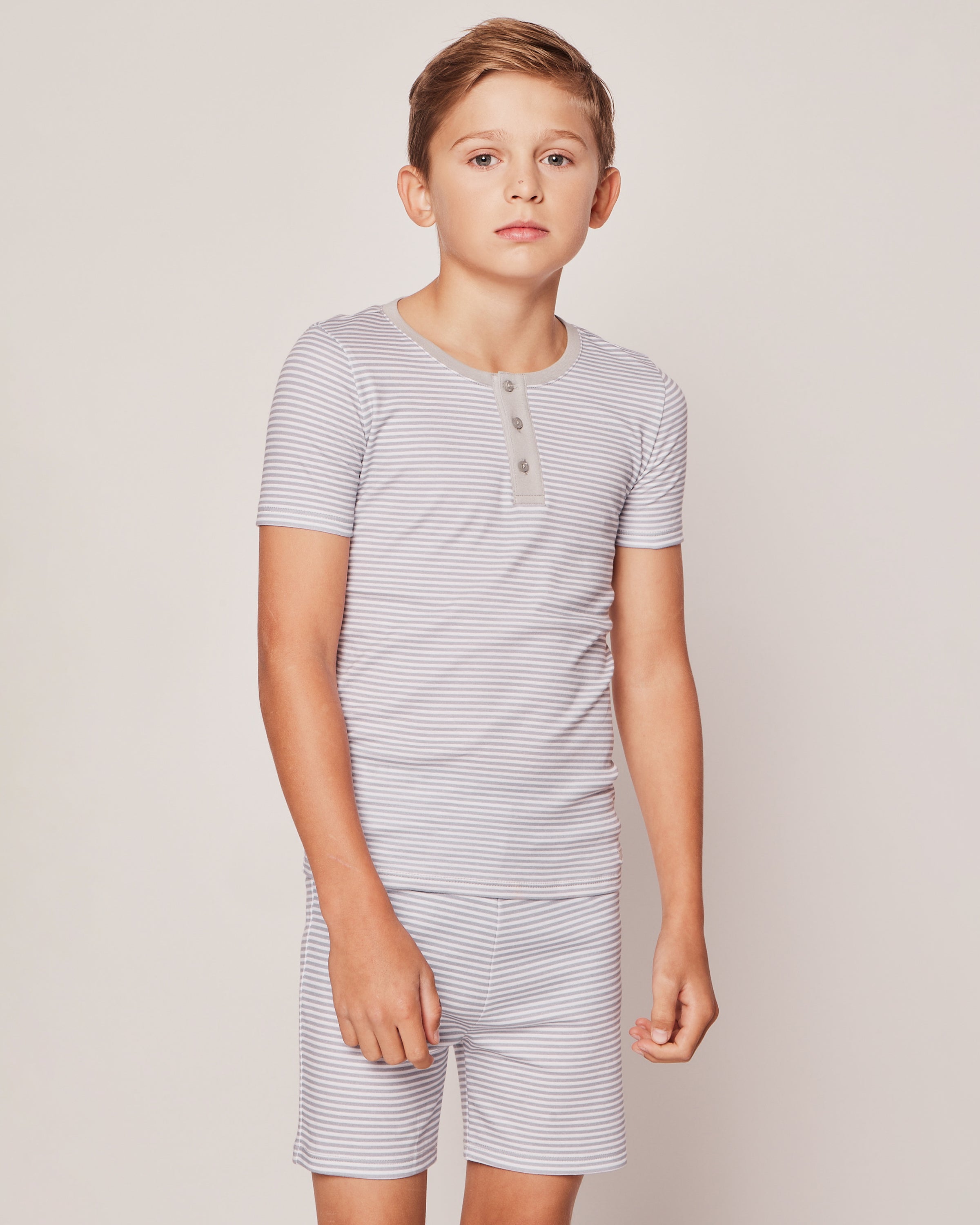 A young boy with short brown hair and a neutral expression stands against a plain background wearing Petite Plumes Kids Pima Snug Fit Pajama Short Set in Grey Stripes, with his right hand slightly raised.