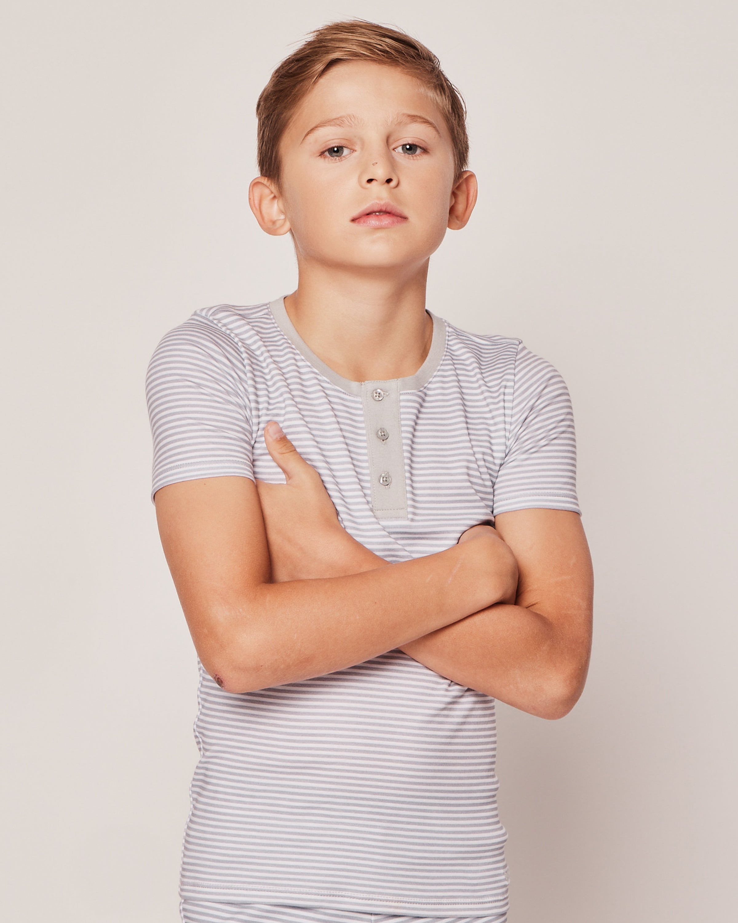 A young boy stands confidently with arms crossed, wearing the Petite Plume Kids Pima Snug Fit Pajama Short Set in Grey Stripes. The background is plain and light-colored.