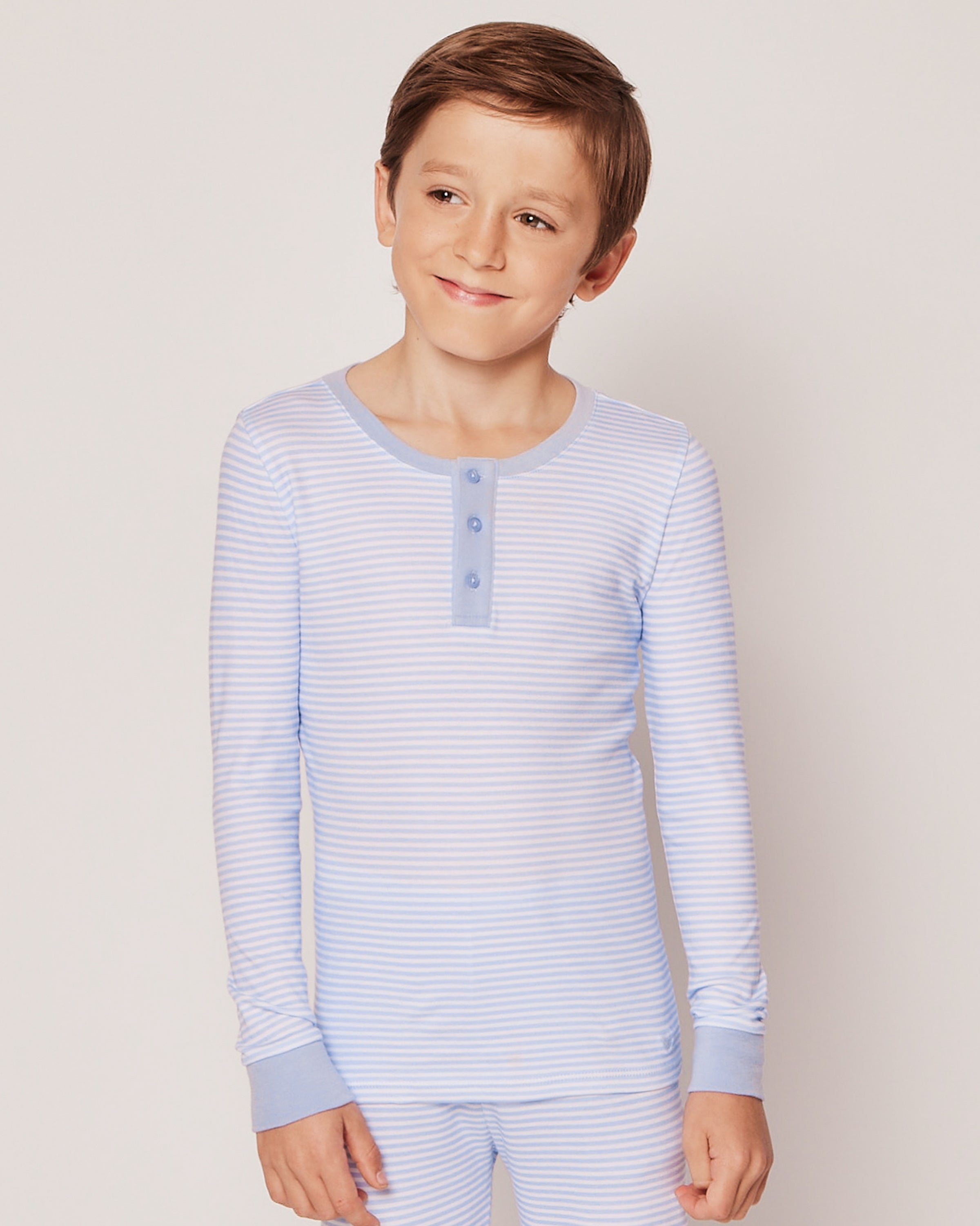 A young boy with short brown hair wears the Kids Pima Snug Fit Pajama Set in Blue Stripes by Petite Plume. The button-up top and matching long pants offer comfort and style as he smiles against a plain light background.