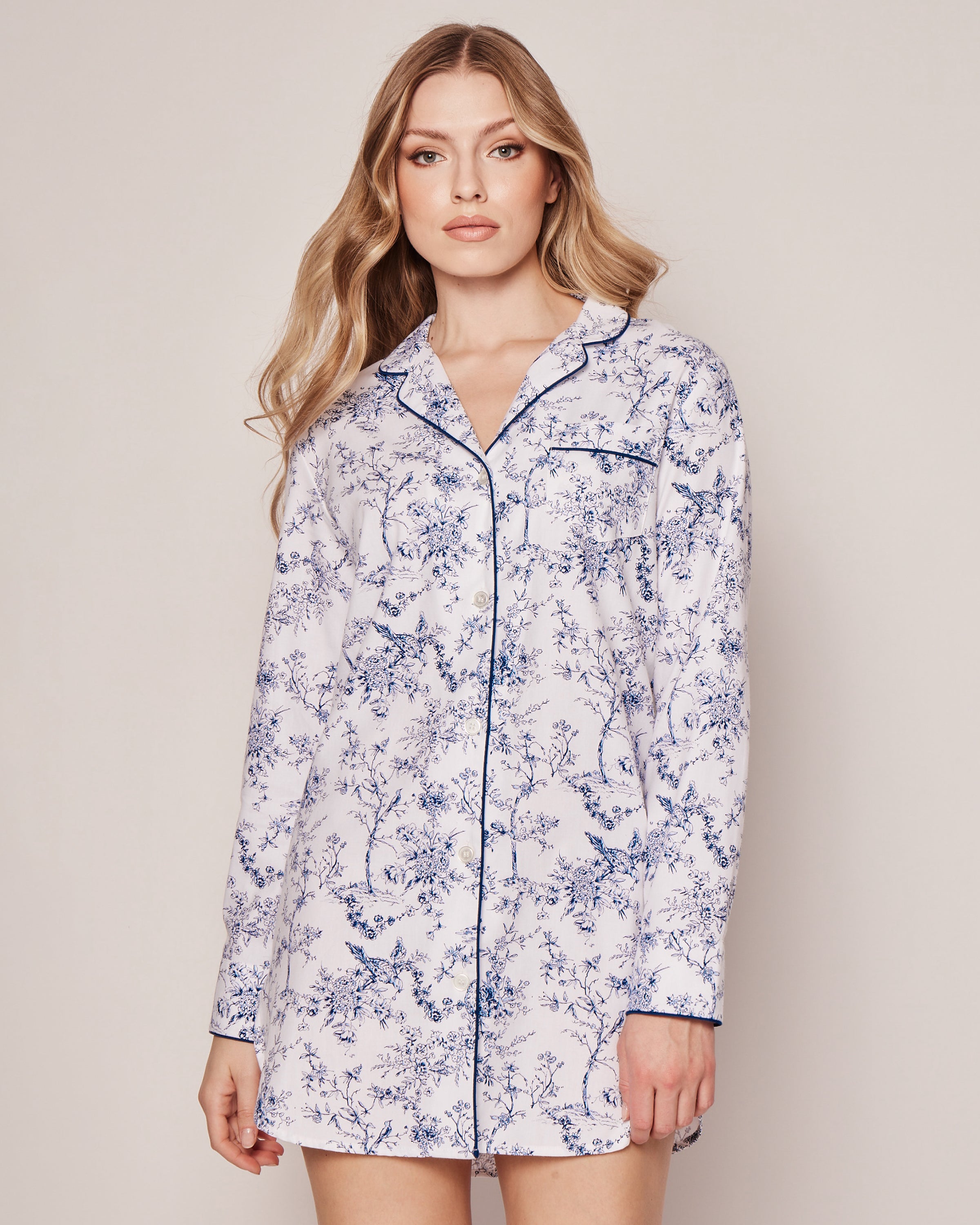 Against a plain backdrop, a person with long, wavy hair wears Petite Plumes Womens Twill Nightshirt in Timeless Toile. The white nightshirt features blue floral patterns, dark piping along the edges, a button-down front, and is crafted from premium cotton for botanical elegance.