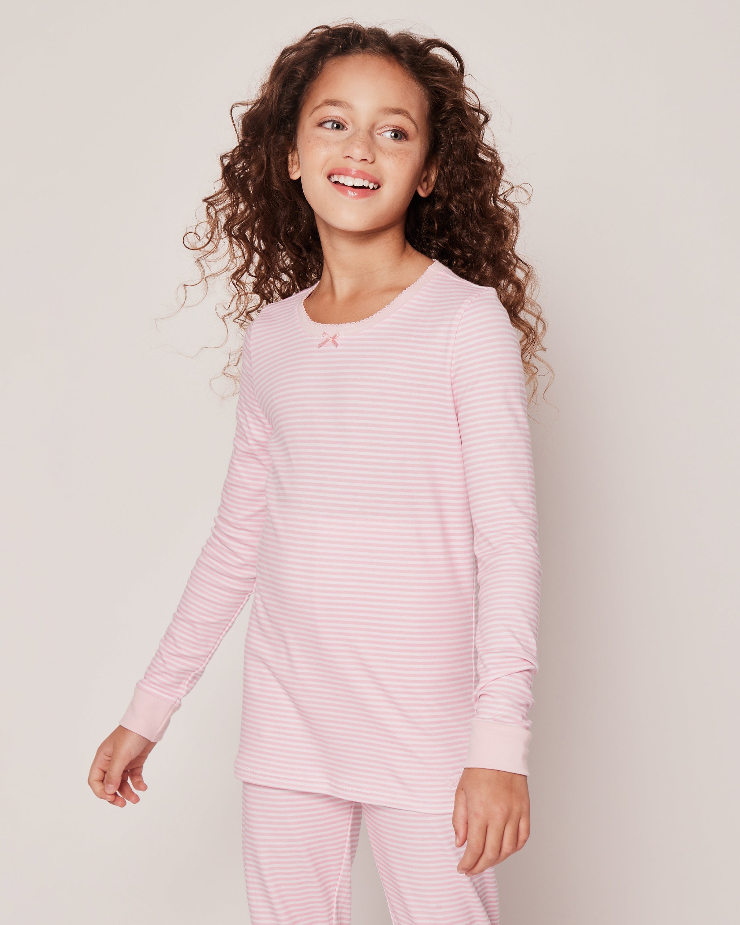A young girl with long curly hair smiles in Petite Plumes Kid’s Pima Snug Fit Pajama Set in Pink Stripes, featuring a soft long-sleeve top and matching pants, ideal for cozy nights. She stands against a plain white background.