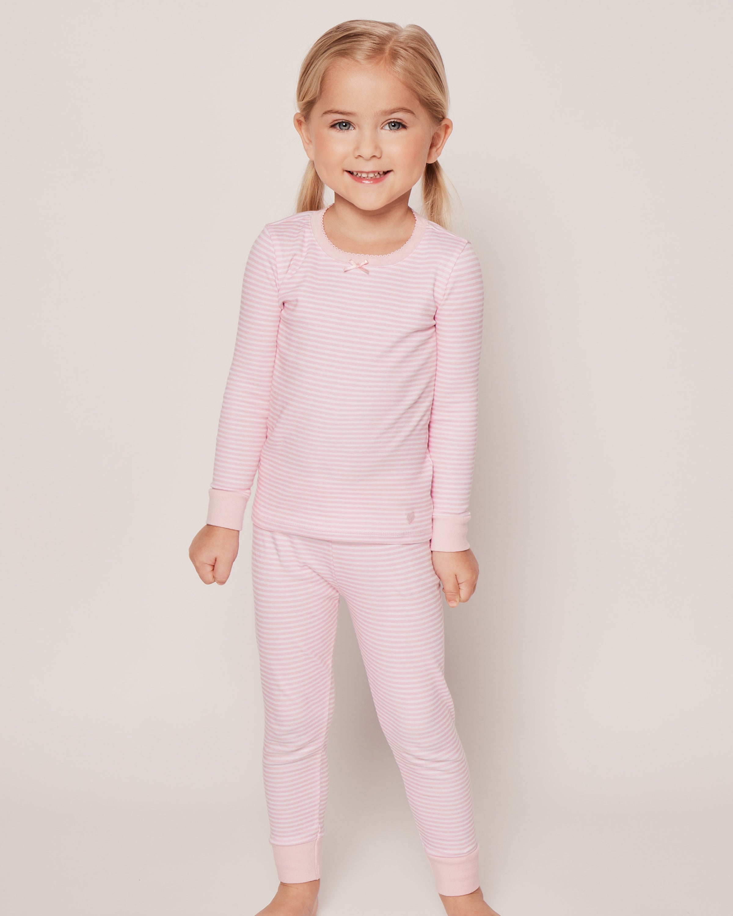 A young child with light hair smiles while wearing Petite Plumes Kids Pima Snug Fit Pajama Set in pink stripes. The cozy, soft Pima cotton and long sleeve top ensure comfort as they stand barefoot against a white background.