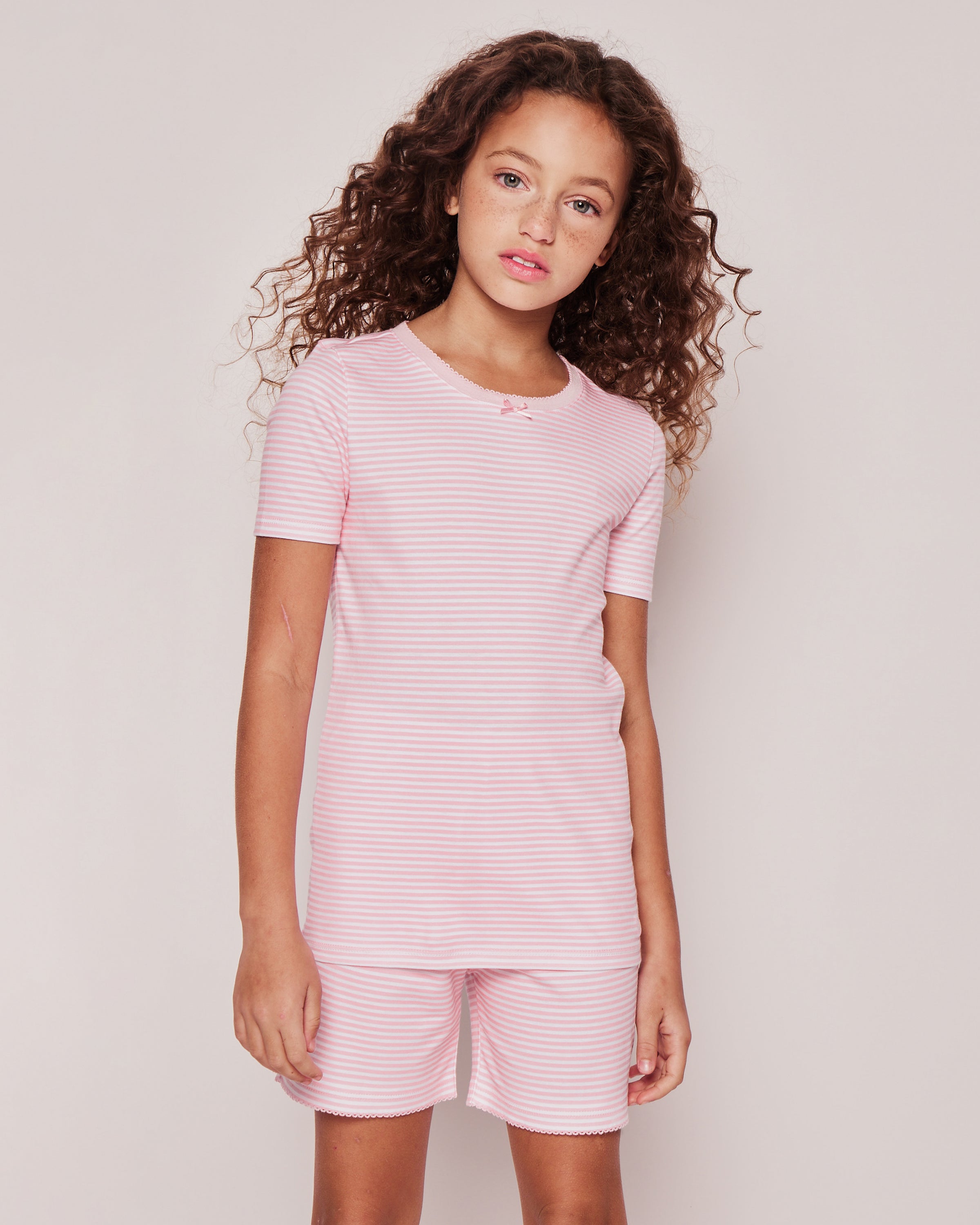 A young girl with curly hair wears the Petite Plume Kids Pima Snug Fit Pajama Short Set in Pink Stripes, gazing neutrally at the camera against a light background, highlighting her adorable sleepwear look.