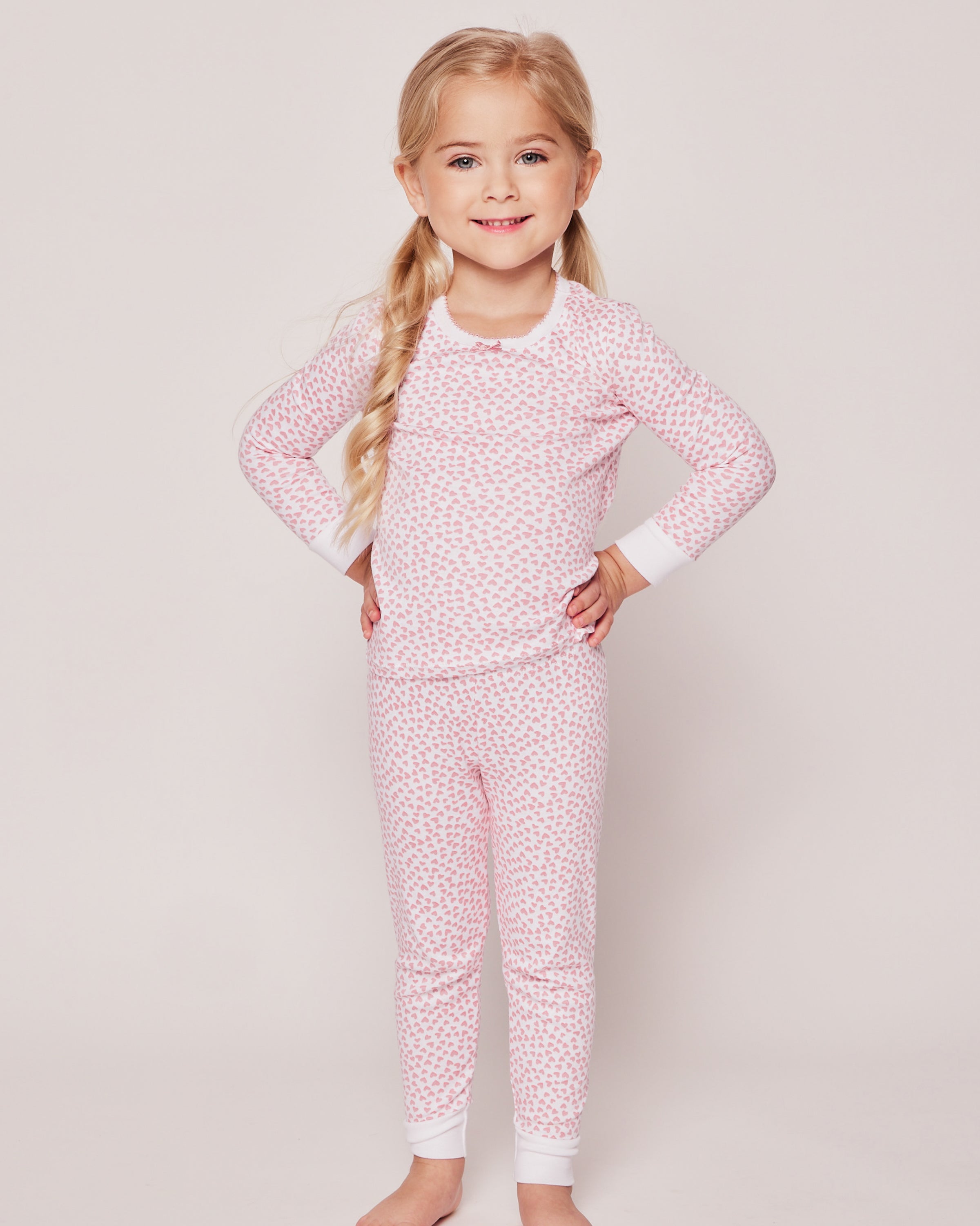 A young girl with long blond hair smiles in Petite Plumes Girls Pima Snug Fit Pajama Trio, featuring a pink and white long sleeve top with matching pants, shown barefoot against a plain white background.
