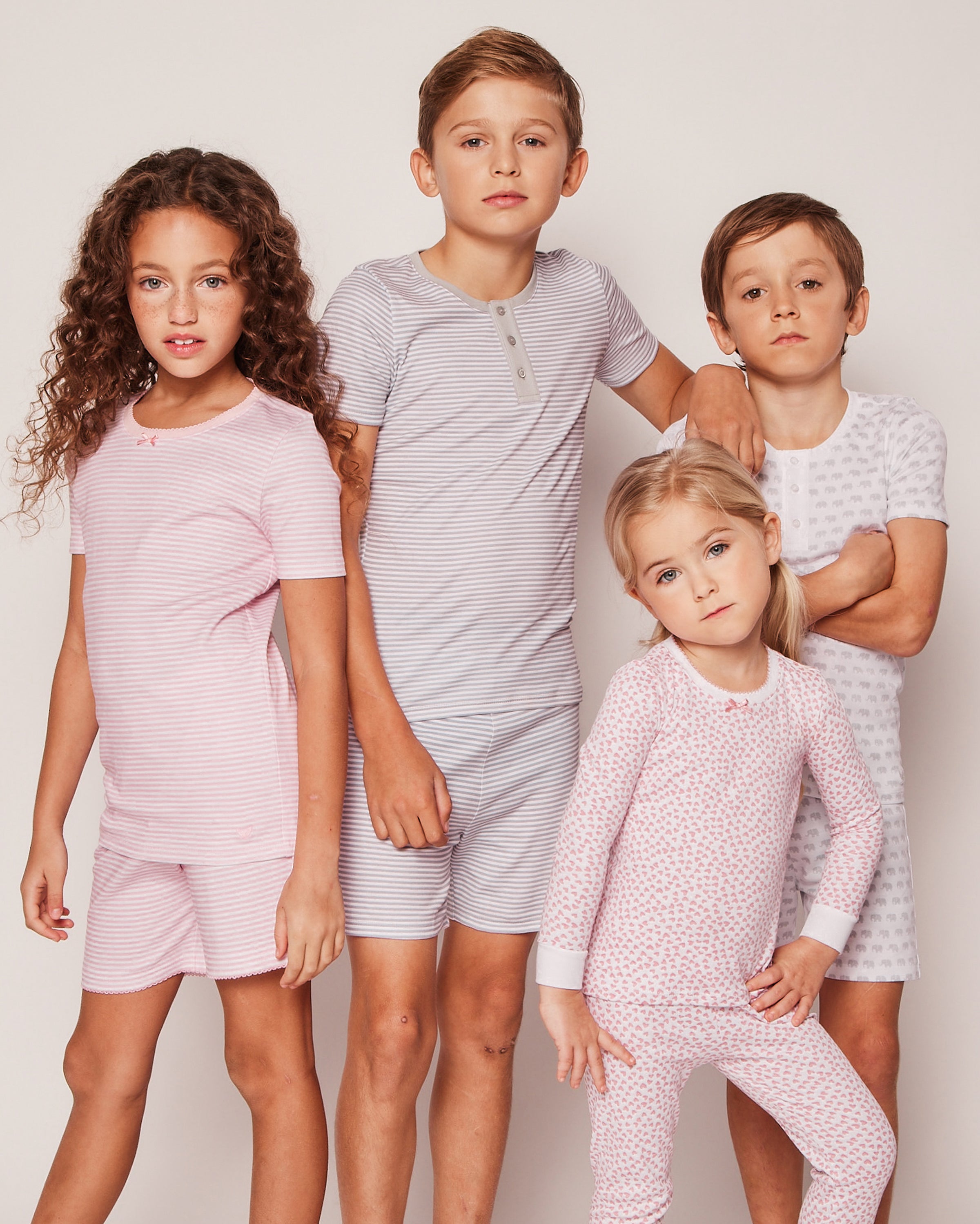 Four children pose with neutral expressions against a plain background, wearing matching Petite Plume Kids Pima Snug Fit Pajama Short Set in Grey Stripes. The cozy sleepwear features Pima cotton, short sleeves, and combines striped and small patterned designs in pastel shades.