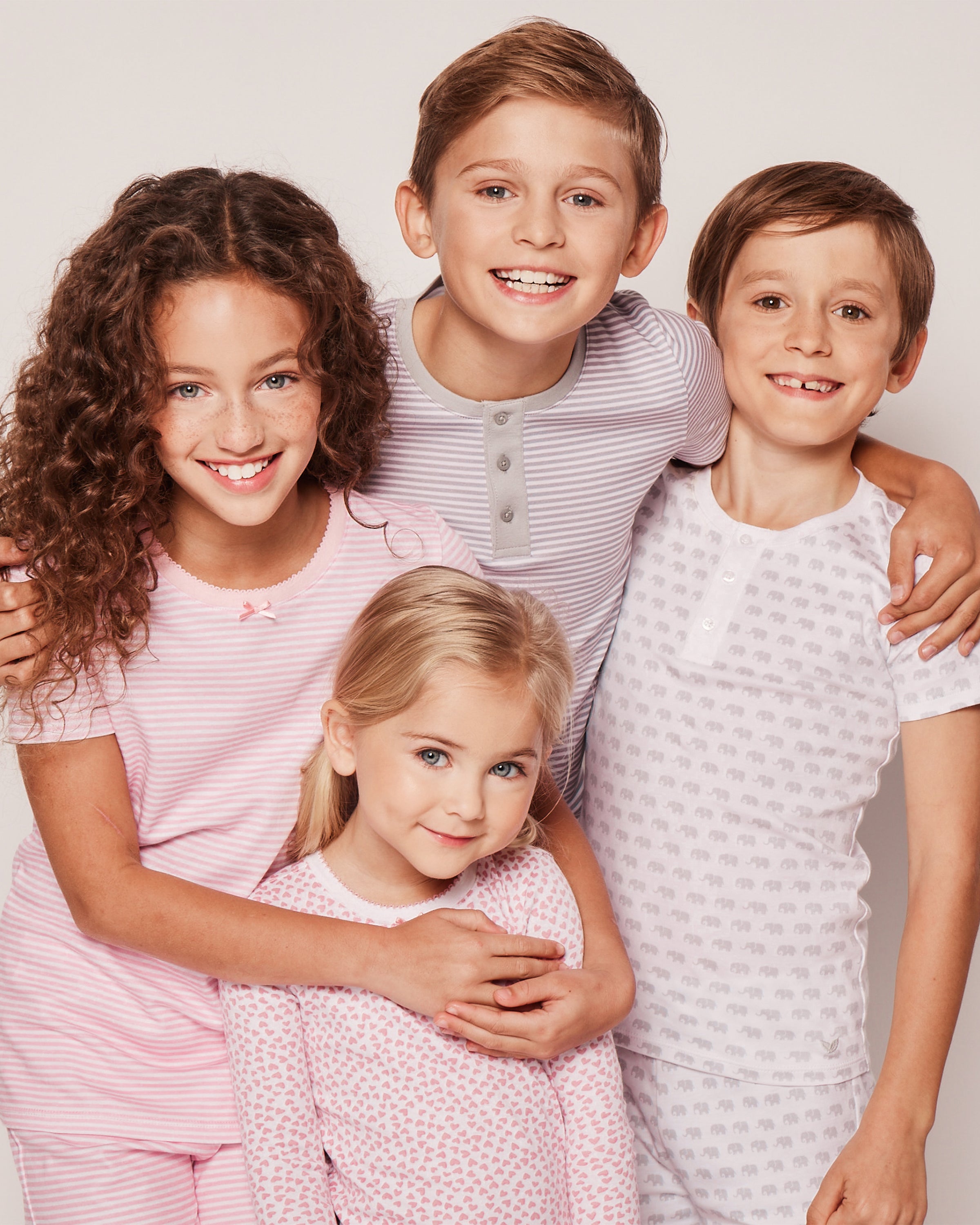 Four children, dressed in Petite Plume luxury sleepwear, smile for the camera. One wears the Kids Pima Snug Fit Pajama Short Set in Grey Elephants, set against a plain background with their arms around each other conveying warmth and togetherness in 100% Pima Cotton comfort.