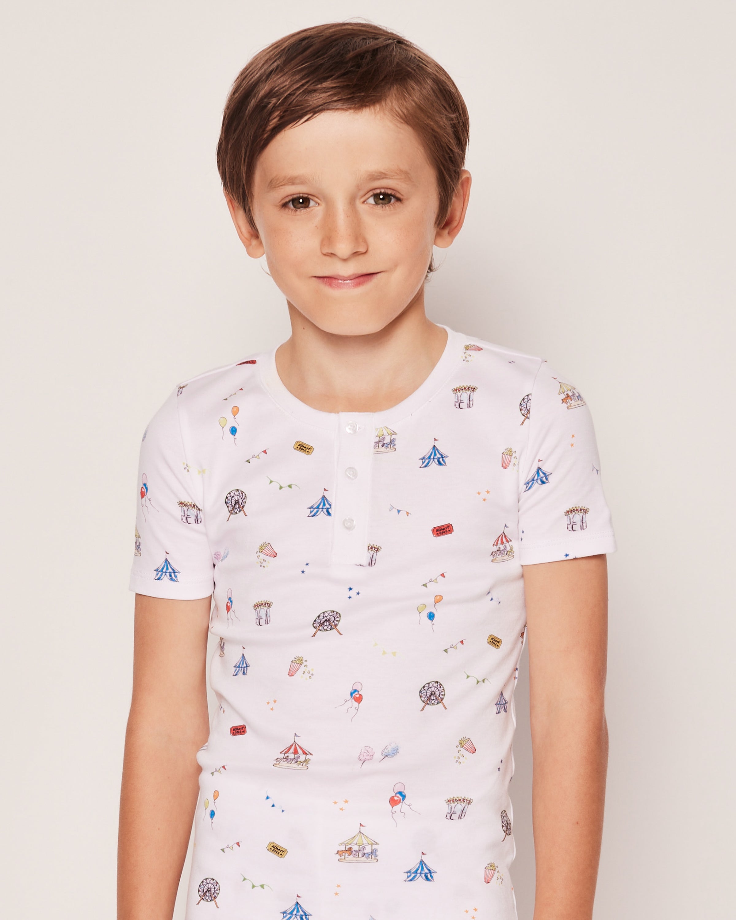 A young boy with short brown hair smiles while wearing Petite Plumes Kids Pima Snug Fit Pajama Short Set in Carnival Memories, featuring small colorful illustrations like balloons and Ferris wheels. The soft fabric ensures comfort as he stands against a plain background.