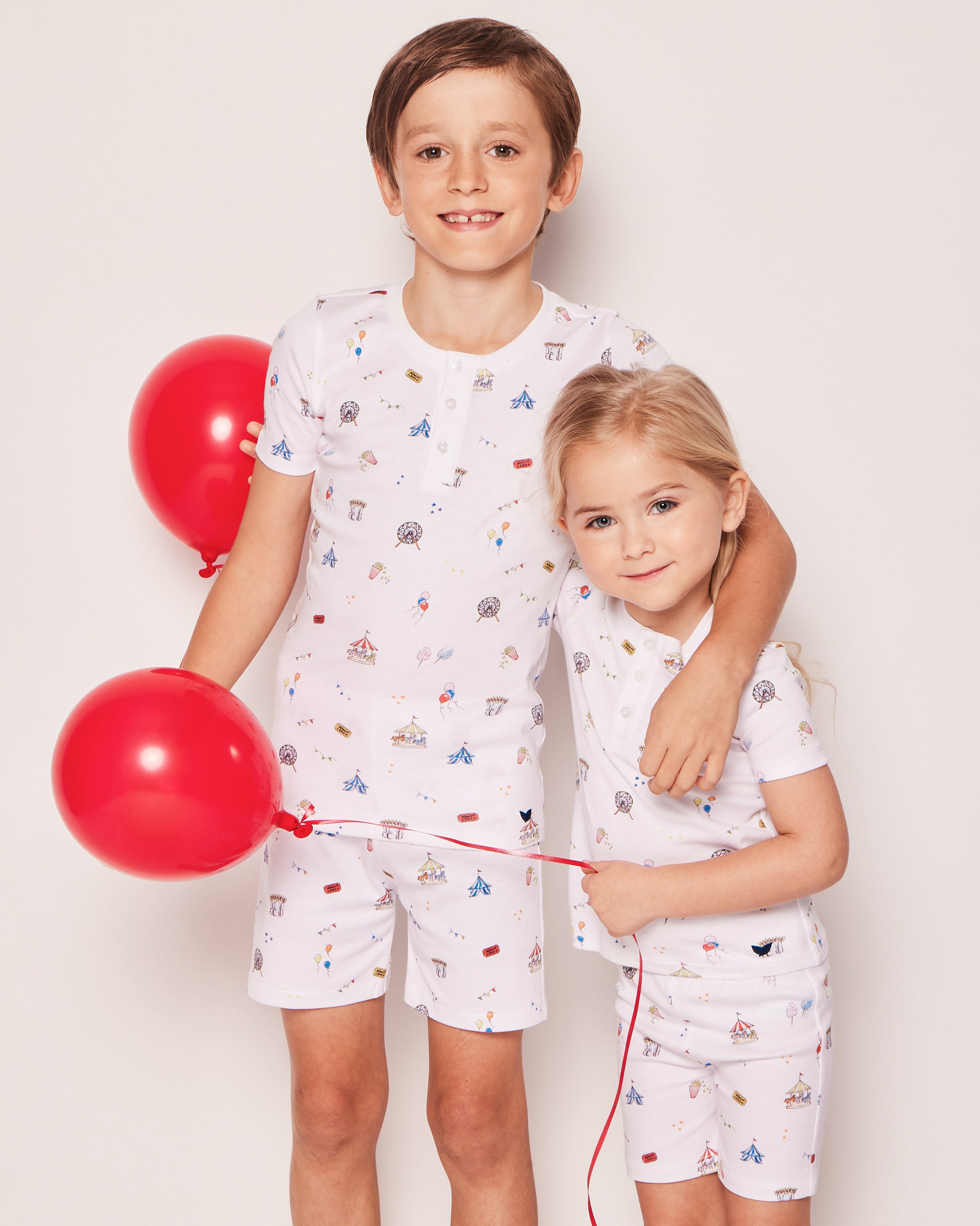 Two kids, each holding red balloons, smile and stand together in Petite Plumes Kids Pima Snug Fit Pajama Short Set in Carnival Memories against a plain white background.