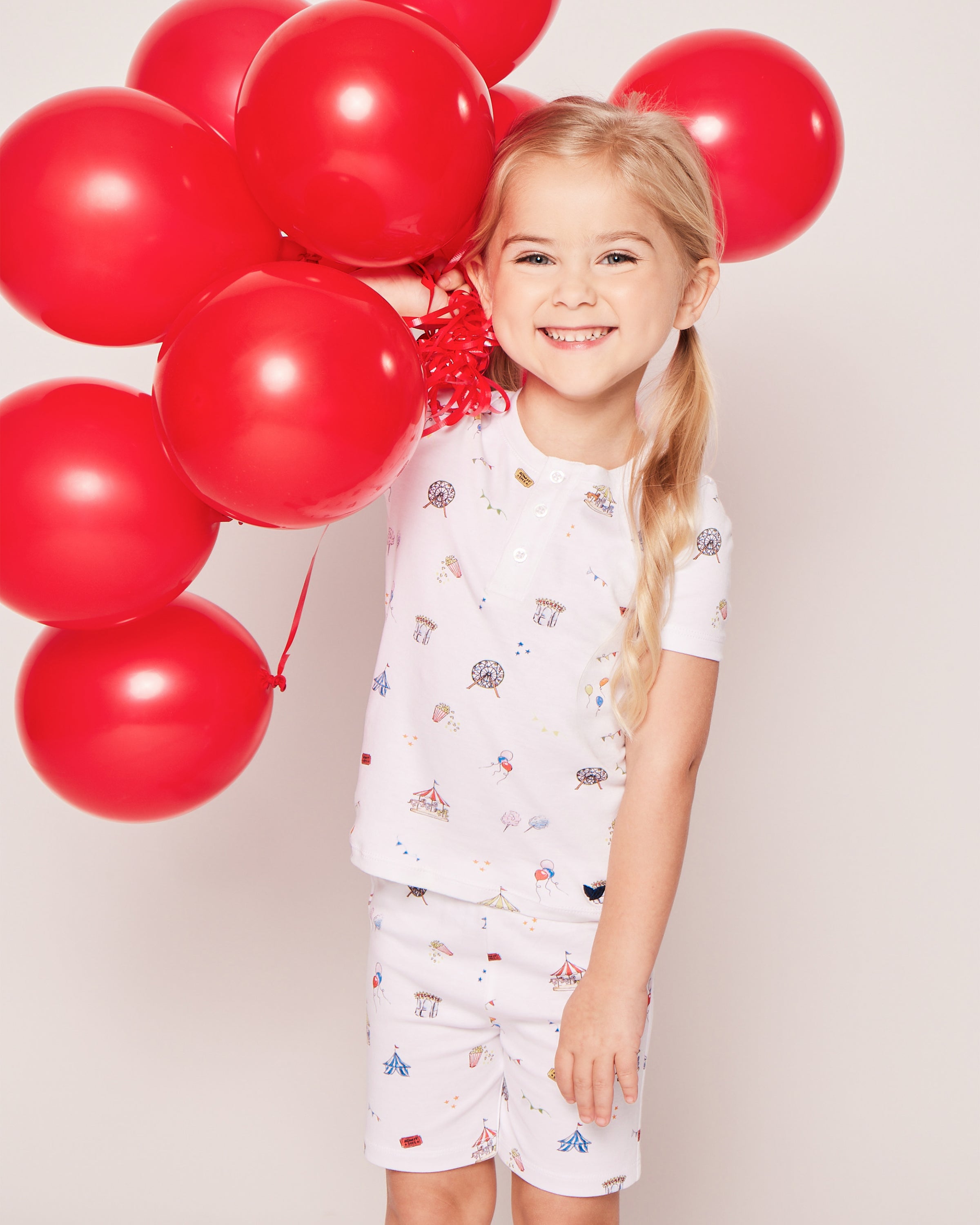 A cheerful blonde girl holds red balloons, wearing Petite Plumes Kids Pima Snug Fit Pajama Short Set in Carnival Memories. The plain, light background accentuates her joyful presence.