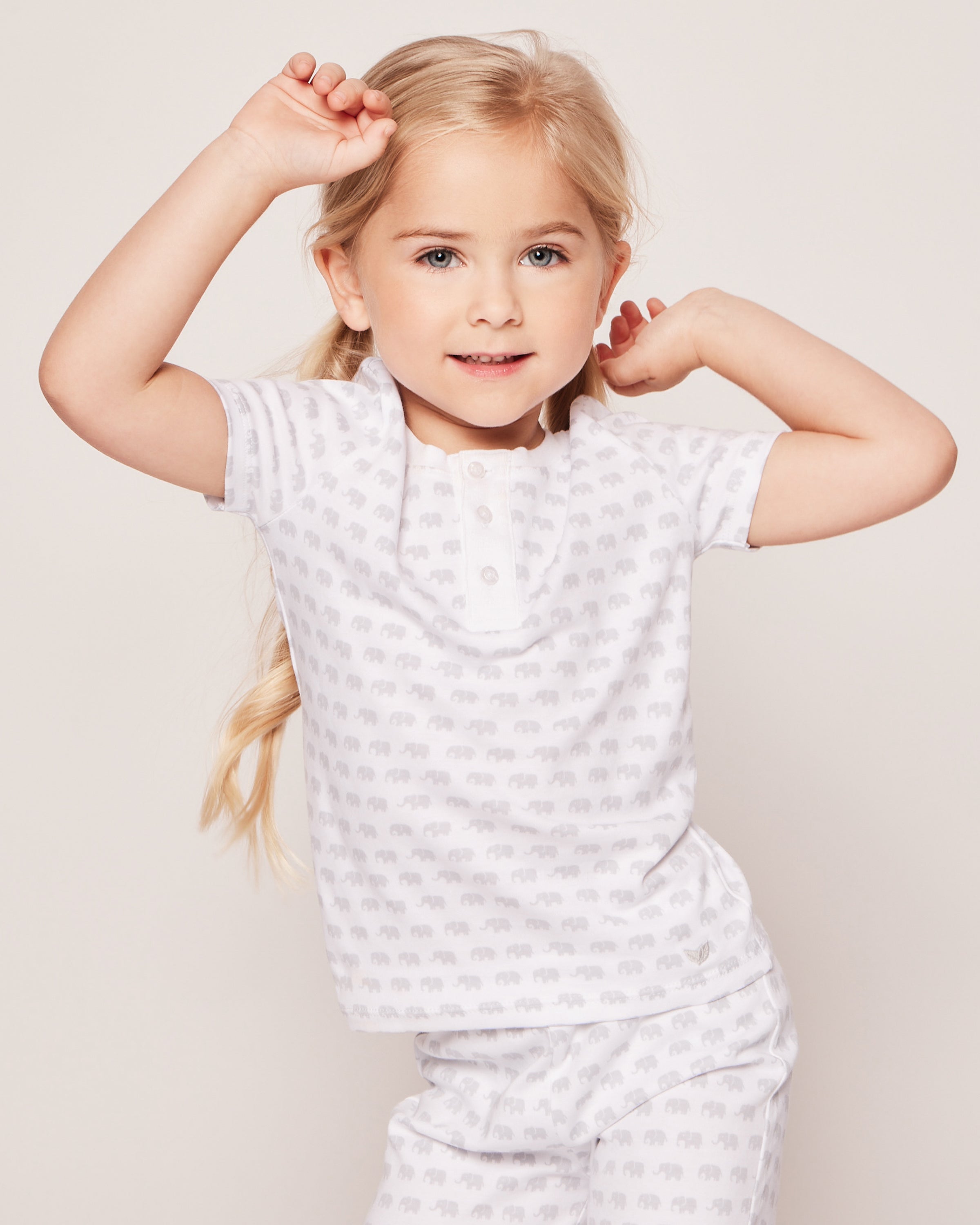 A young girl with long blonde hair confidently poses in Petite Plumes Kids Pima Snug Fit Pajama Short Set in Grey Elephants. Her white pajamas with a grey elephants print complement her playful smile as she raises her arms against a plain, light background.