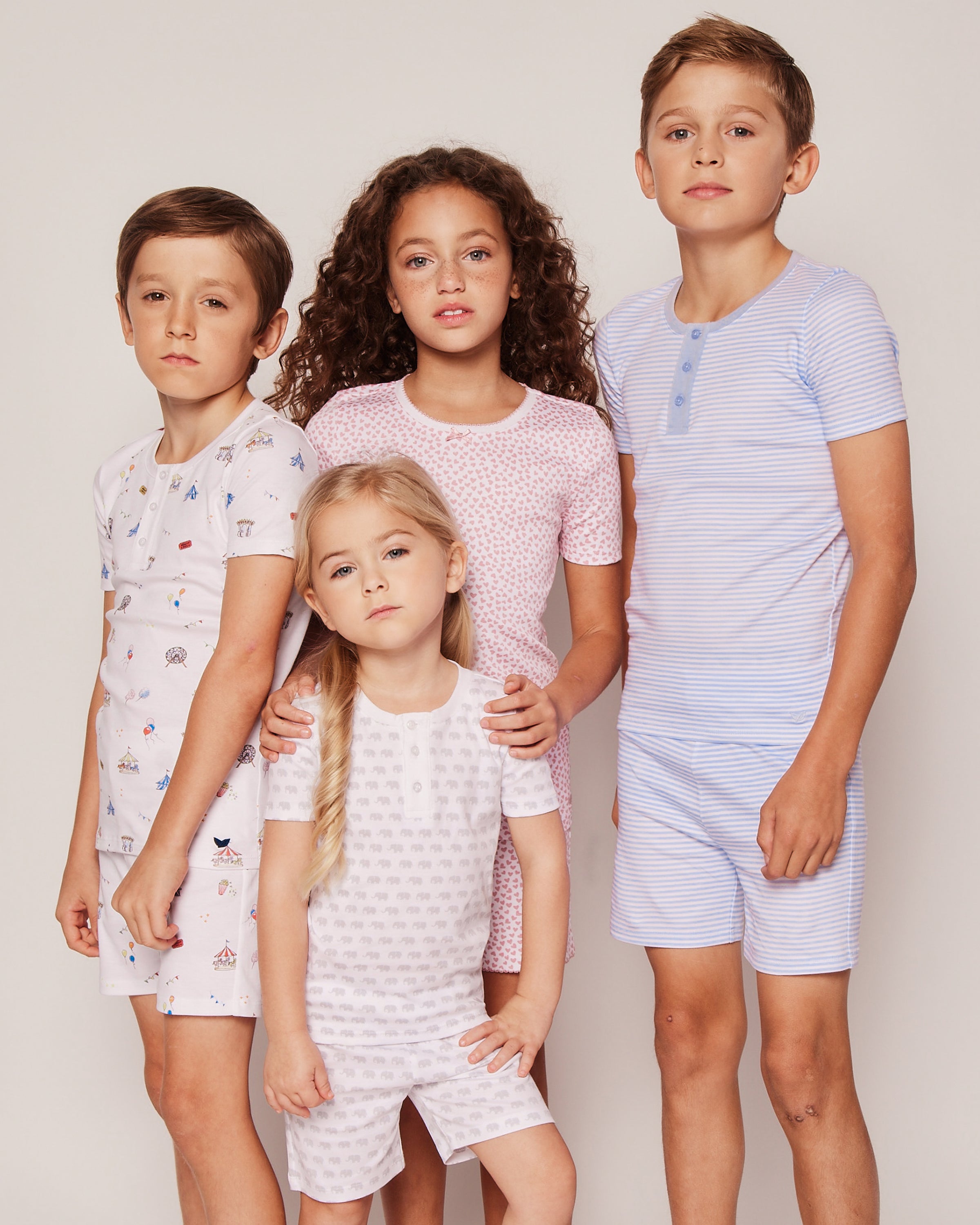 Four kids in cozy sleepwear stand against a neutral backdrop. Three boys wear striped and patterned pajamas, while a girl with curls joins them. Another young girl wears the Kids Pima Snug Fit Pajama Short Set in Carnival Memories by Petite Plume.