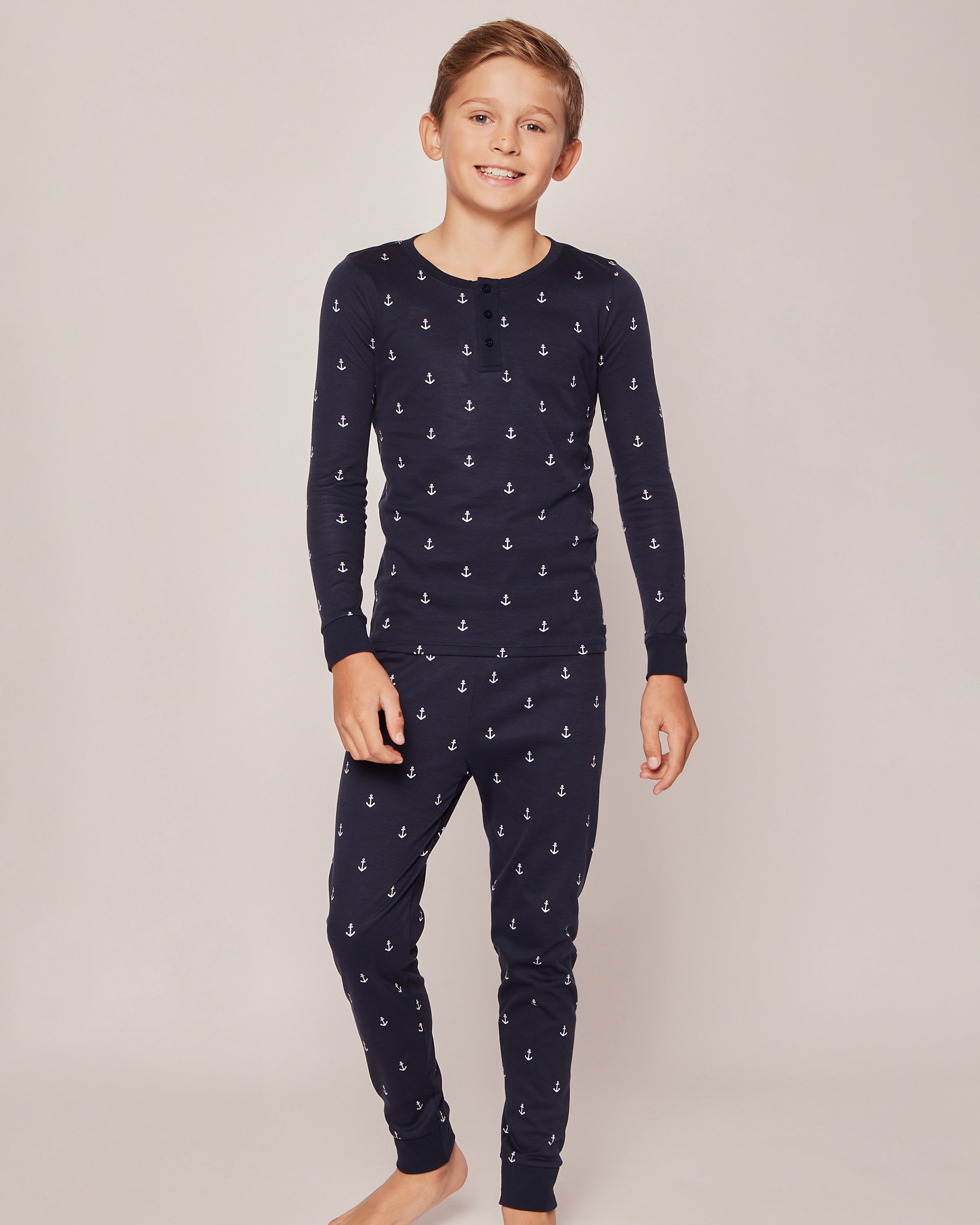 A young boy beams in his cozy Petite Plume Kids Pima Snug Fit Pajama Set in Portsmouth Anchors, featuring a navy blue design with small white anchor patterns. He stands barefoot against a plain light grey background, dressed in the long sleeve top and pant set.
