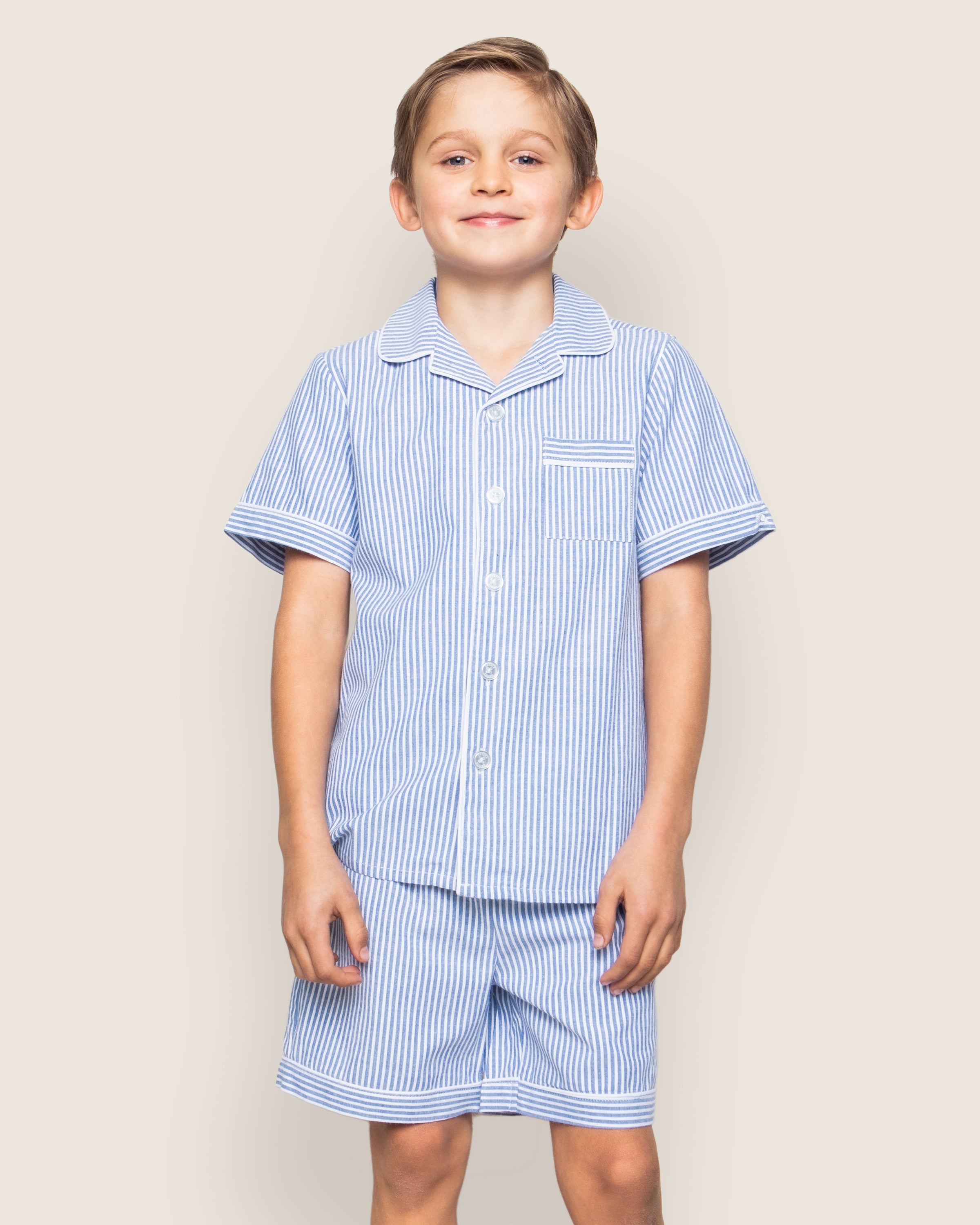 A young boy smiles in Petite Plumes Kids Twill Pajama Short Set, featuring short sleeves and a collar. Made from lightweight cotton, the French Blue Seersucker set includes a chest pocket and matching shorts. He stands against a plain light beige background.