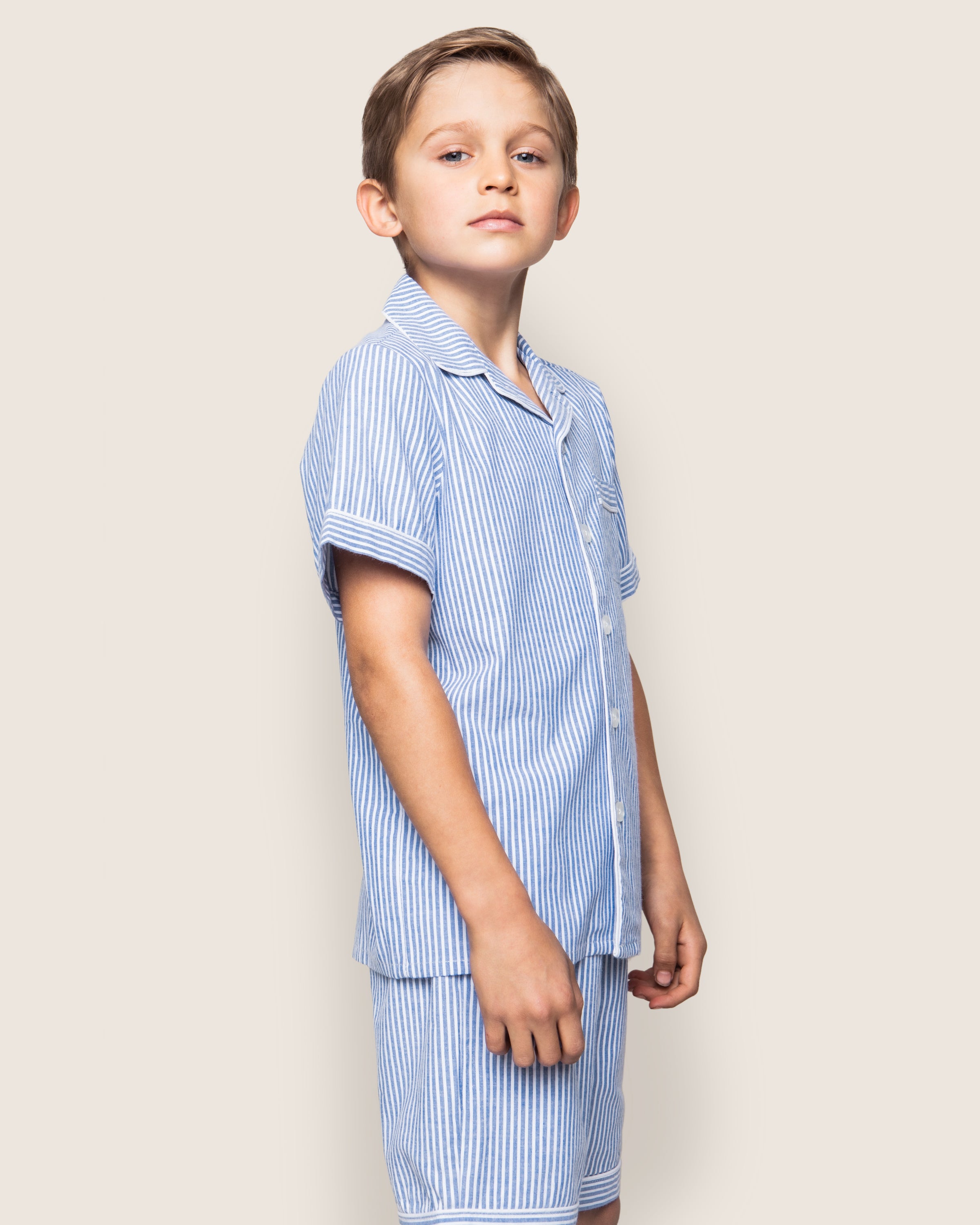A young boy stands confidently in the Kids Twill Pajama Short Set in French Blue Seersucker from Petite Plume, crafted from lightweight cotton. The short-sleeve button-up shirt and shorts ensemble complements his neutral expression as he gazes slightly upwards against a plain beige background.