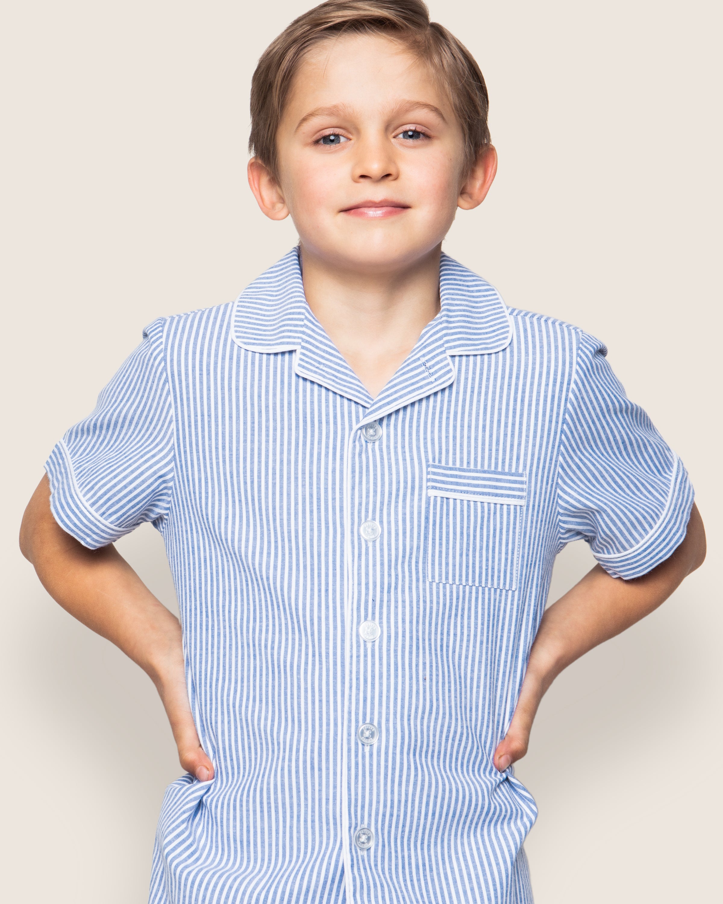 A short-haired child smiles confidently with hands on hips, wearing Petite Plumes Kids Twill Pajama Short Set in French Blue Seersucker, featuring a light blue and white striped shirt with a collar and breast pocket against a plain backdrop.