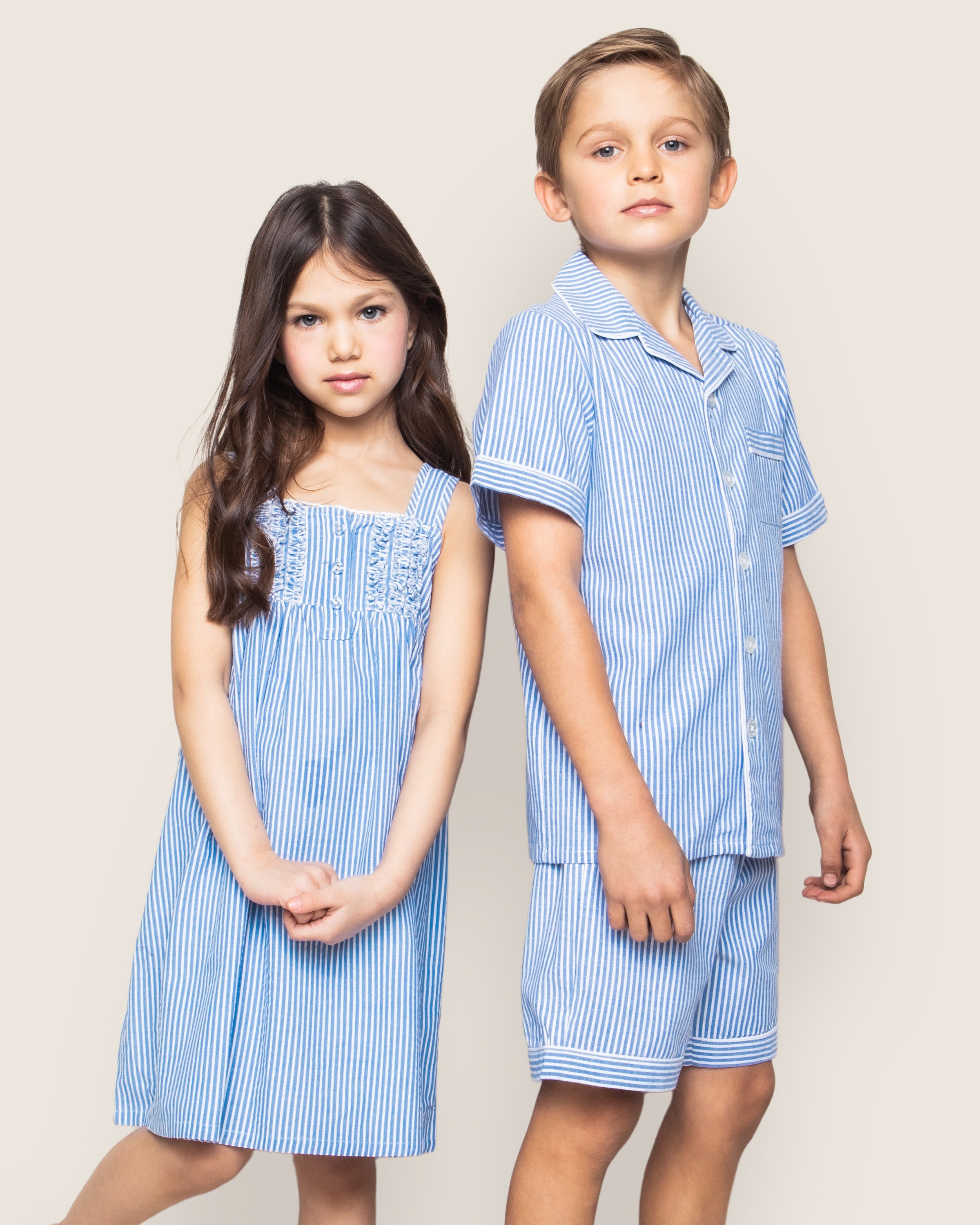A young girl in a blue and white striped dress stands beside a boy in Petite Plumes Kids Twill Pajama Short Set in French Blue Seersucker. Their neutral expressions complement the plain backdrop as they showcase this classic yet cozy sleepwear.