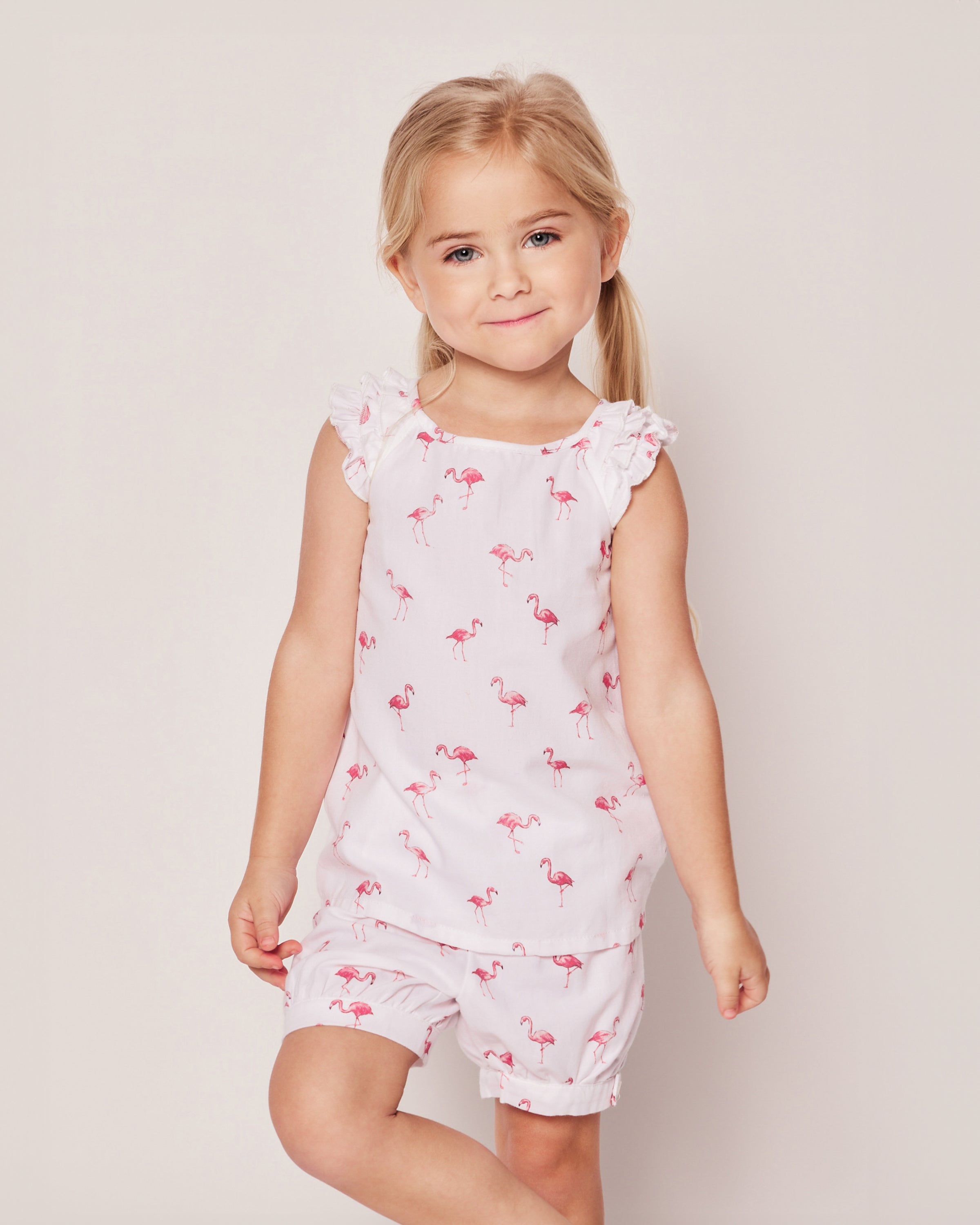 A young blonde girl wears the Girls Twill Amelie Short Set in Flamingos by Petite Plume, featuring a sleeveless top with ruffled shoulders and matching shorts in a playful pink flamingo pattern. She smiles gently against a plain, light-colored background.
