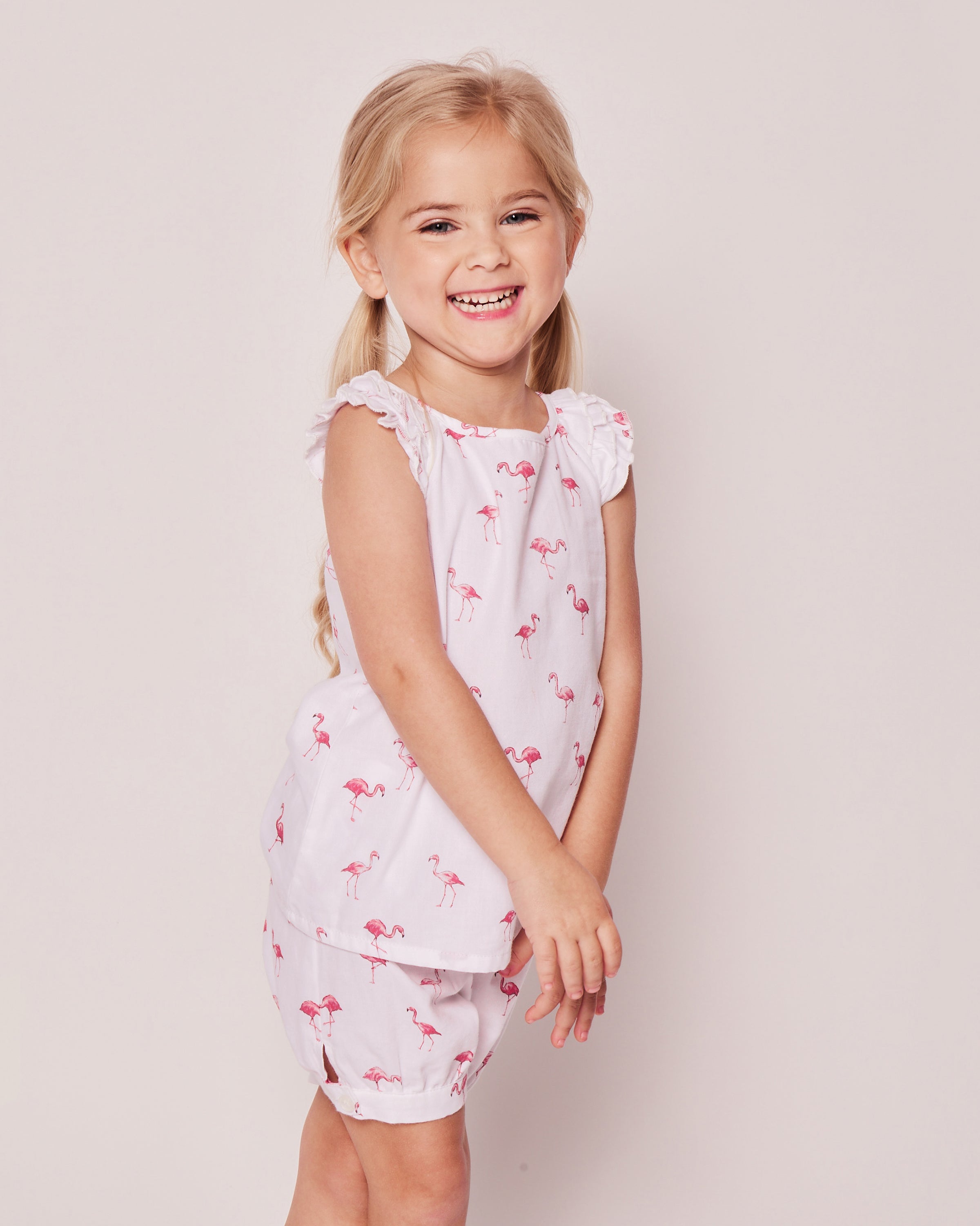 A young girl with blond hair smiles in adorable sleepwear, featuring Petite Plumes Girls Twill Amelie Short Set in Flamingos. Her white sleeveless top and shorts showcase playful pink flamingo patterns, set against a plain, light-colored background.