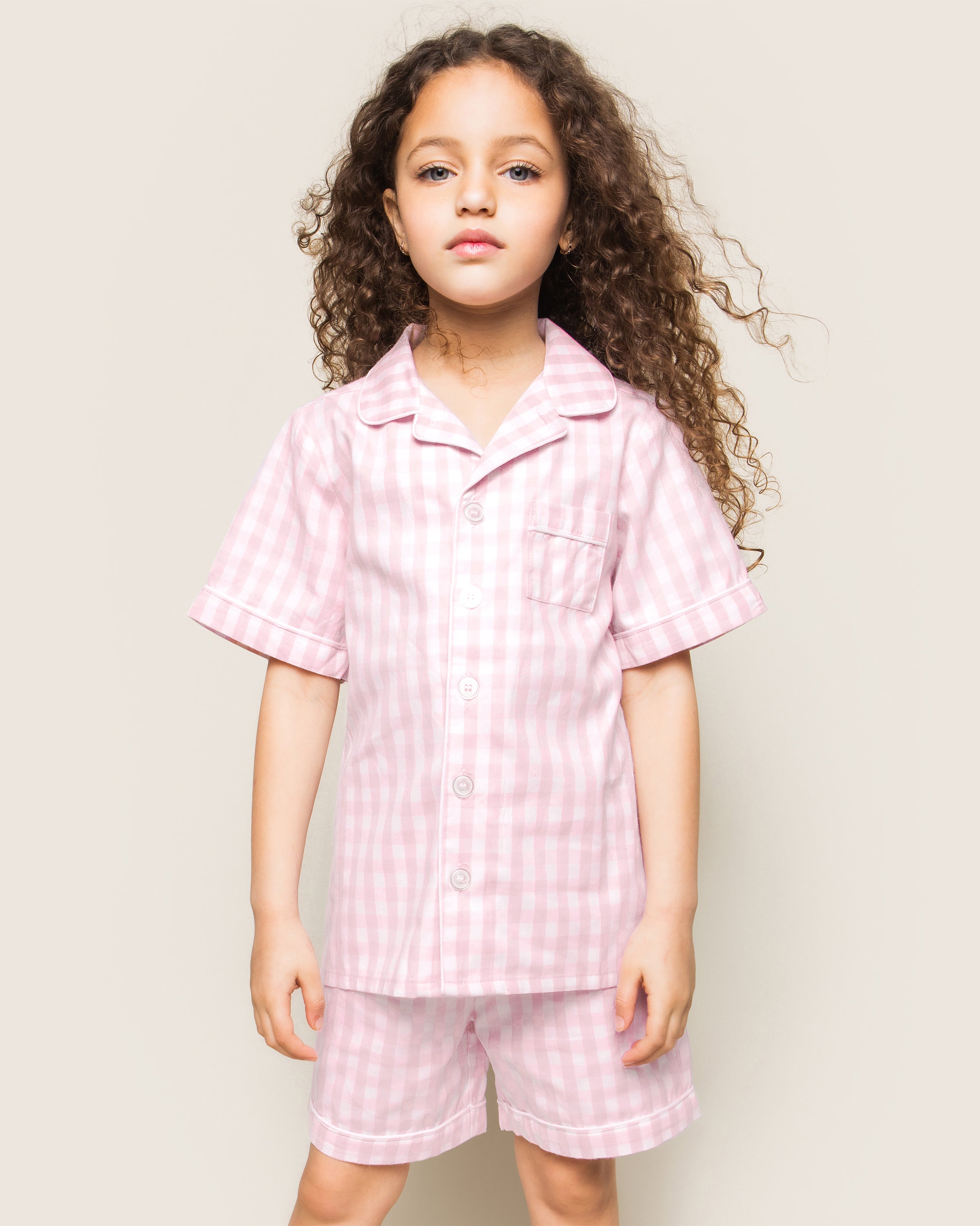 Three folded Petite Plume Kids Twill Pajama Short Sets in pink gingham rest on a textured surface, featuring charming designs of pink hearts, floral toile patterns, and pink stripes. Crafted from yarn-dyed cotton, they exude quality. Elegantly surrounded by white flowers for enhanced presentation.