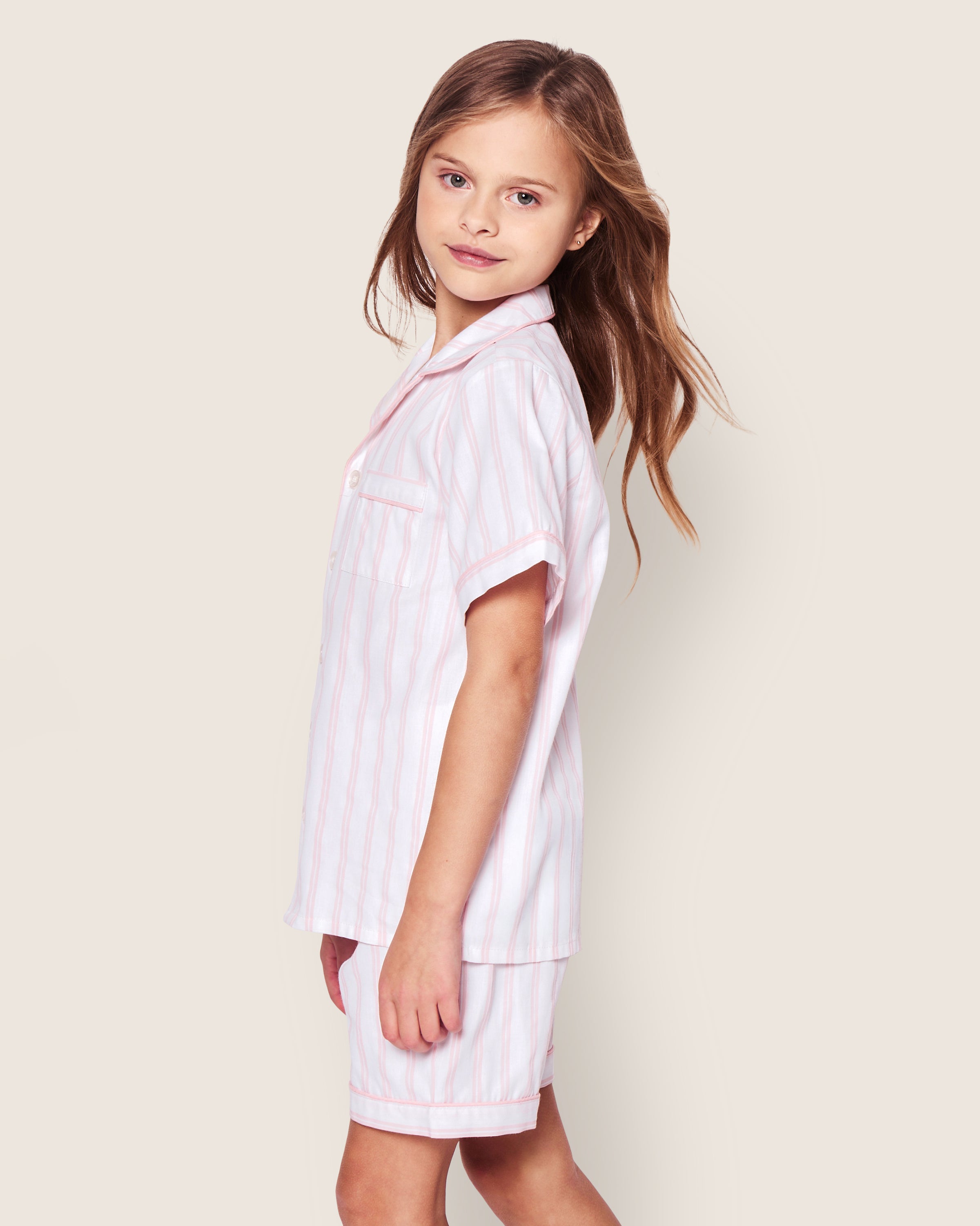 A young girl with long hair stands sideways in a Petite Plume Kids Twill Pajama Short Set in Pink and White Stripe. The soft cotton short-sleeved button-up top and shorts fit her perfectly as she glances back at the camera against a plain beige background.