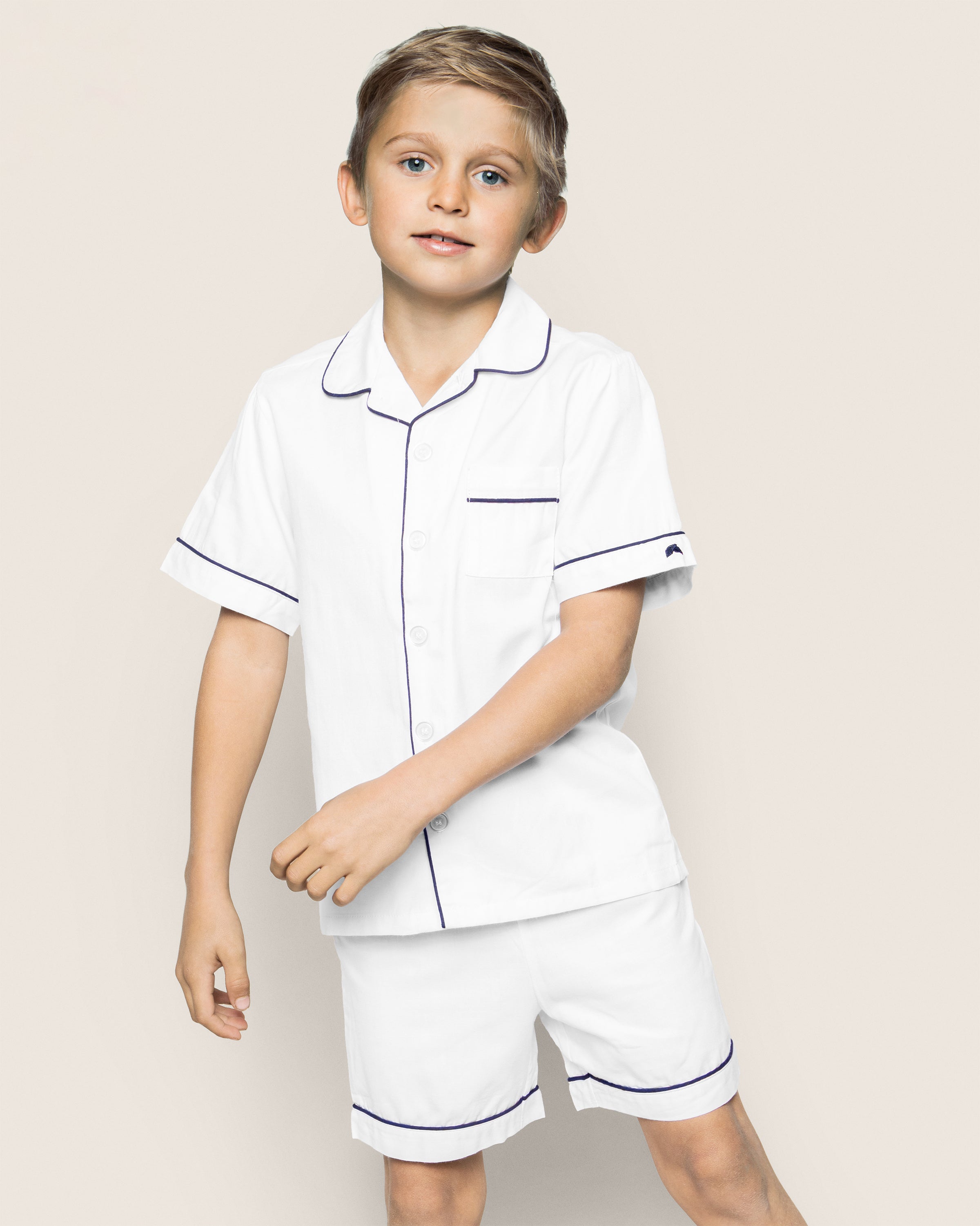 A young boy, with light brown hair and a relaxed expression, stands against a plain background wearing the Kids Twill Pajama Short Set in White with Navy Piping by Petite Plume. The set features soft short sleeves and shorts, capturing the essence of luxurious sleepwear.