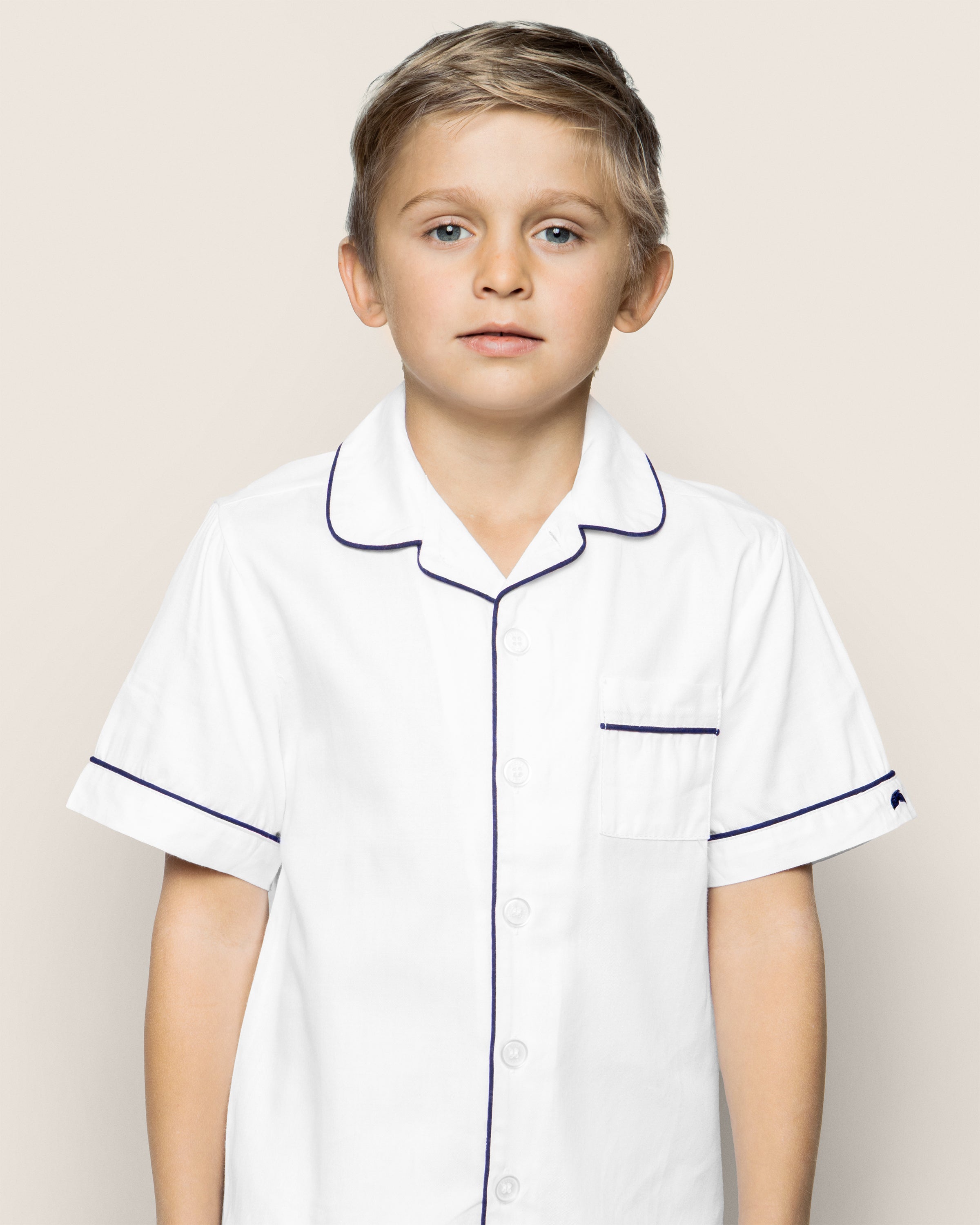 A young boy with short blond hair wears Petite Plumes Kids Twill Pajama Short Set in white with navy piping, standing against a plain beige background and looking straight at the camera with a neutral expression.