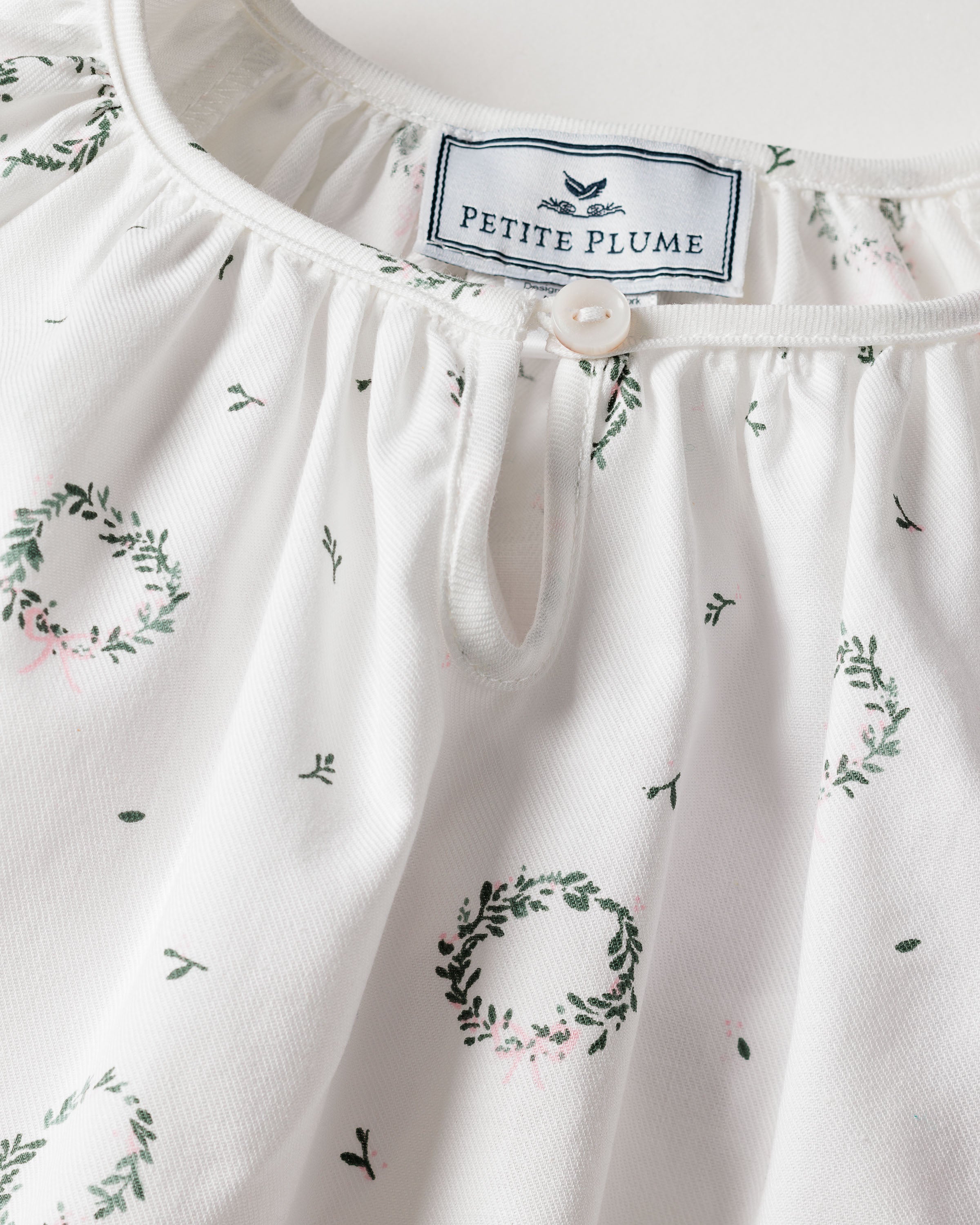 Close-up of a white Petitie Plume garment, the Girls Twill Delphine Nightgown in Somerset Wreath features small green wreath patterns. Made from chemical-free flame-retardant fabric, it includes a label inside near a keyhole button closure at the neckline.