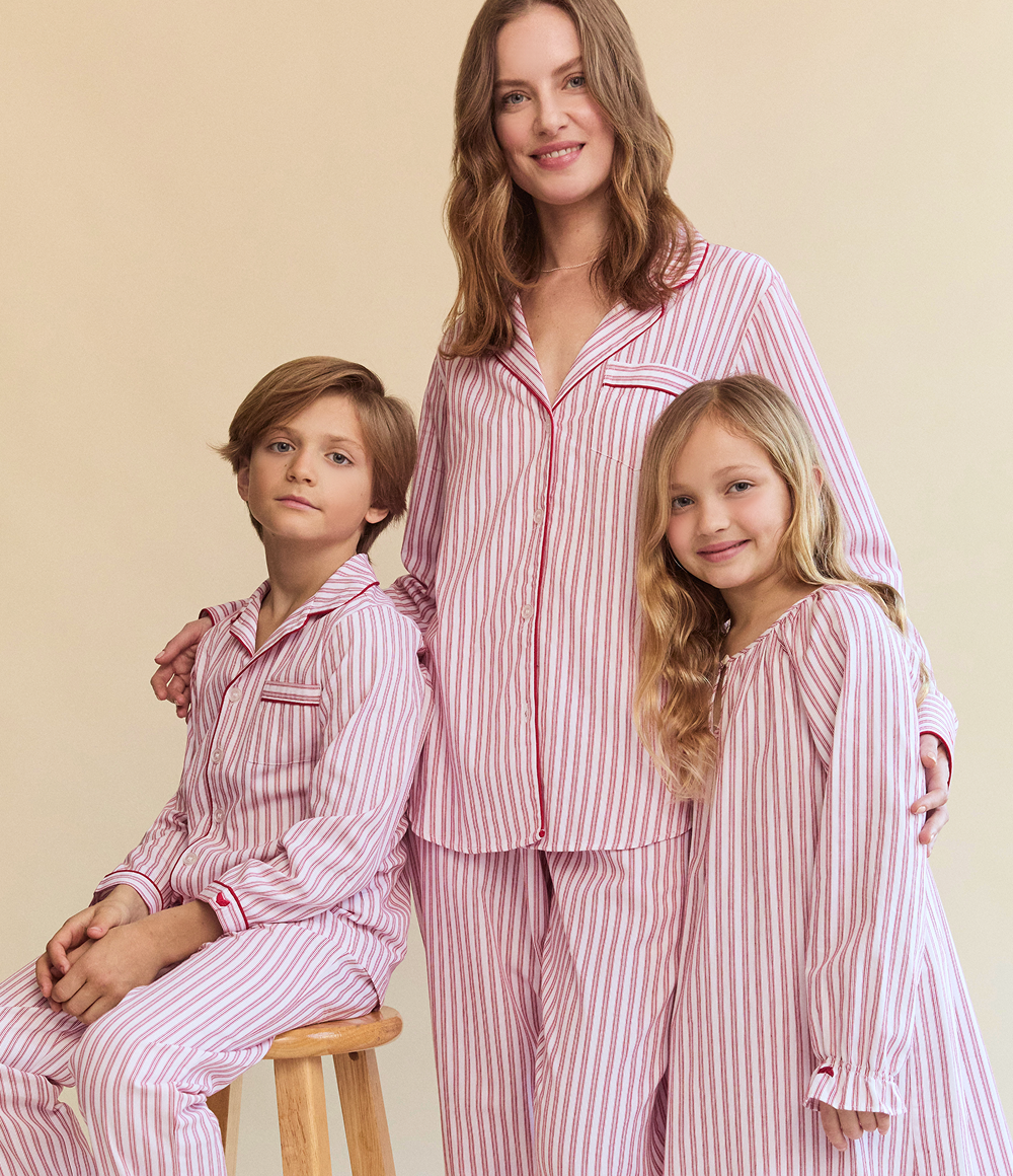 A woman and two children, all wearing matching red and white striped pajamas, pose together. The woman stands behind the two children, who are seated and standing beside her, smiling softly against a plain beige background.